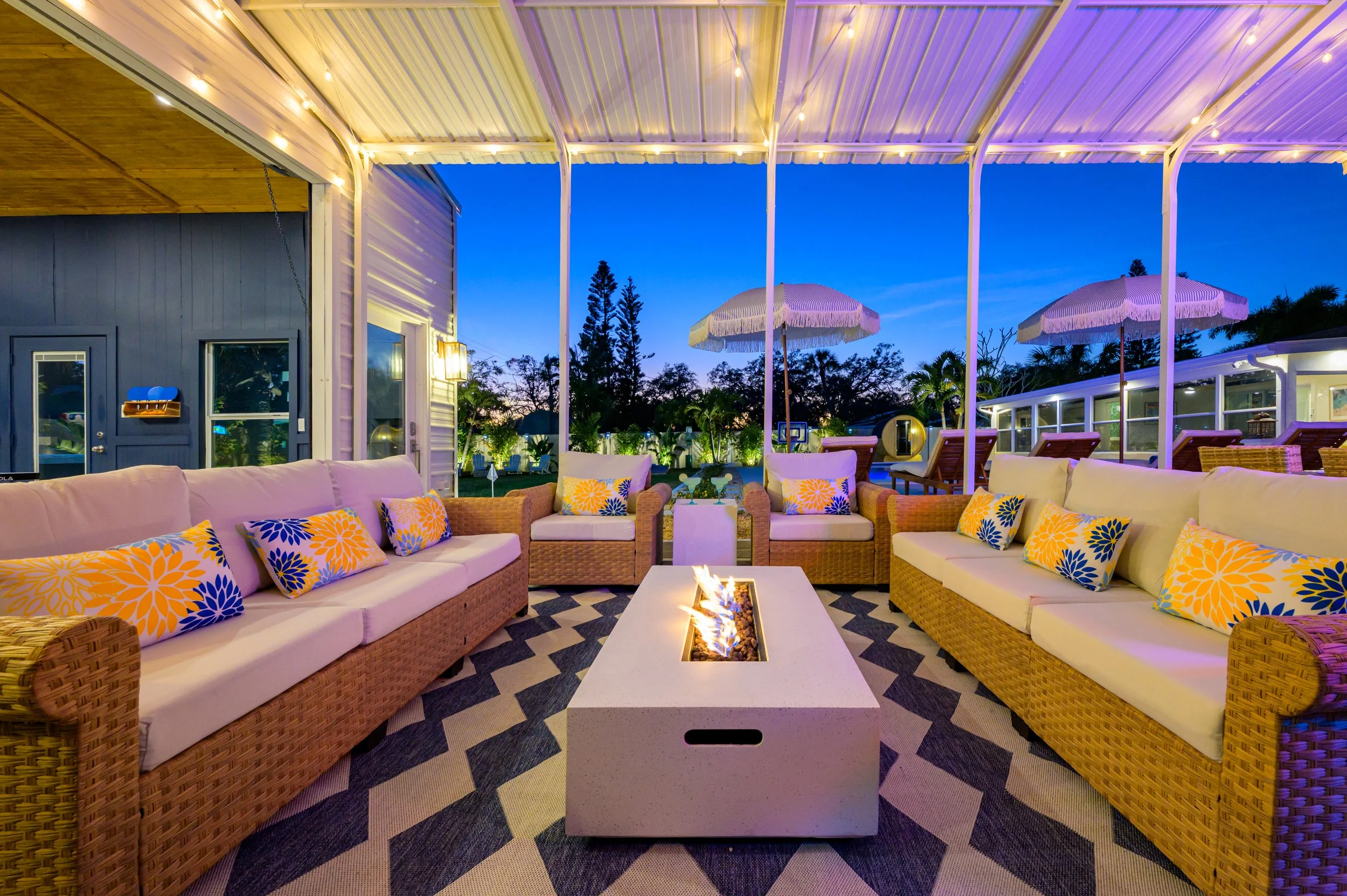 Outdoor patio seating with wicker sofas and colorful pillows, a fire pit table, and string lights, with an evening sky and trees in the background.