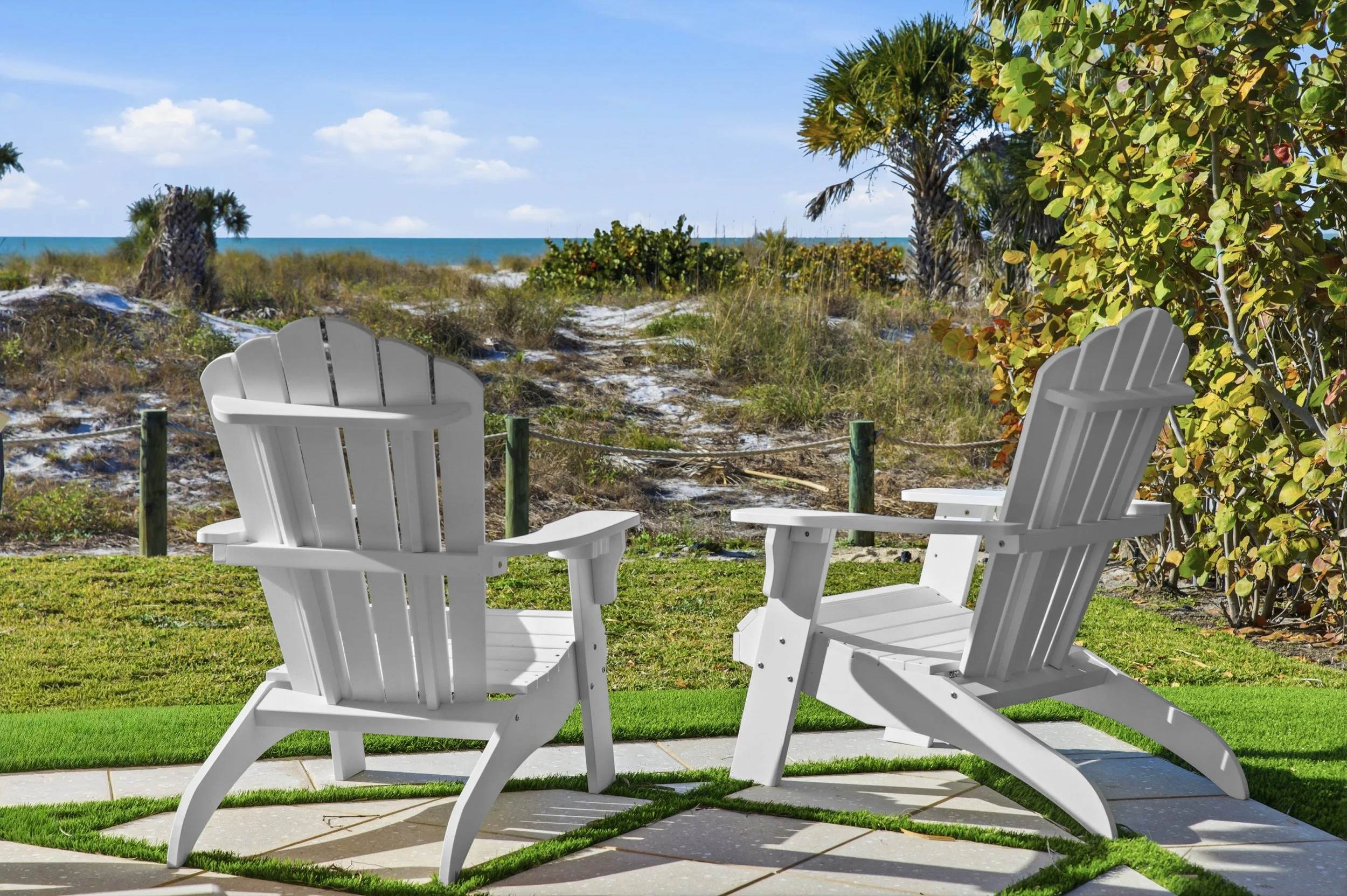 Two white Adirondack chairs on a patio facing a beach with sand dunes, grass, and some bushes, with a clear blue sky and ocean in the background.