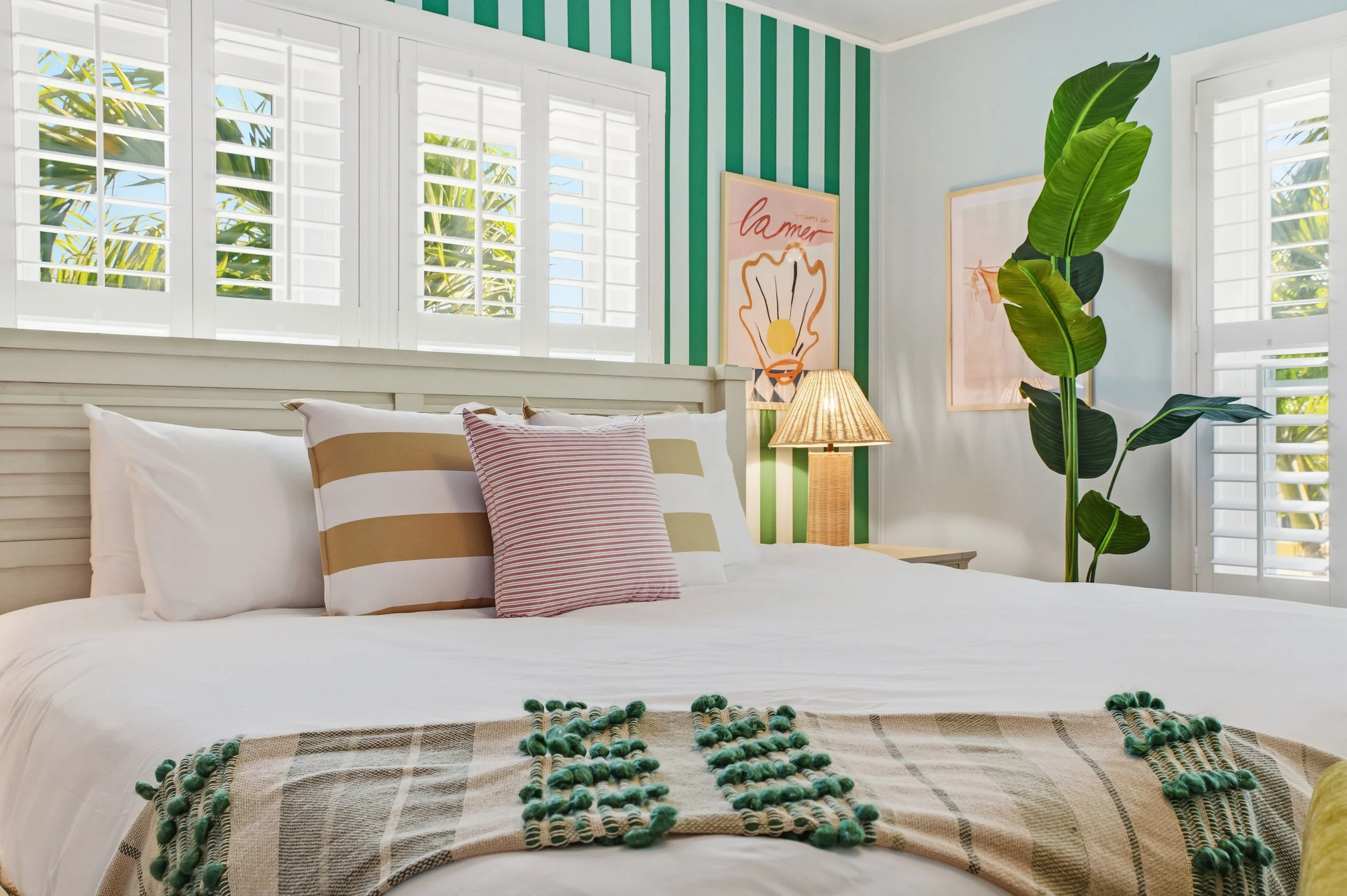 A cozy bedroom with white bedding, decorative pillows, green and beige striped blanket, and large green plant. The room has large windows with white shutters, striped wallpaper on one wall, framed artwork, and a bedside lamp.