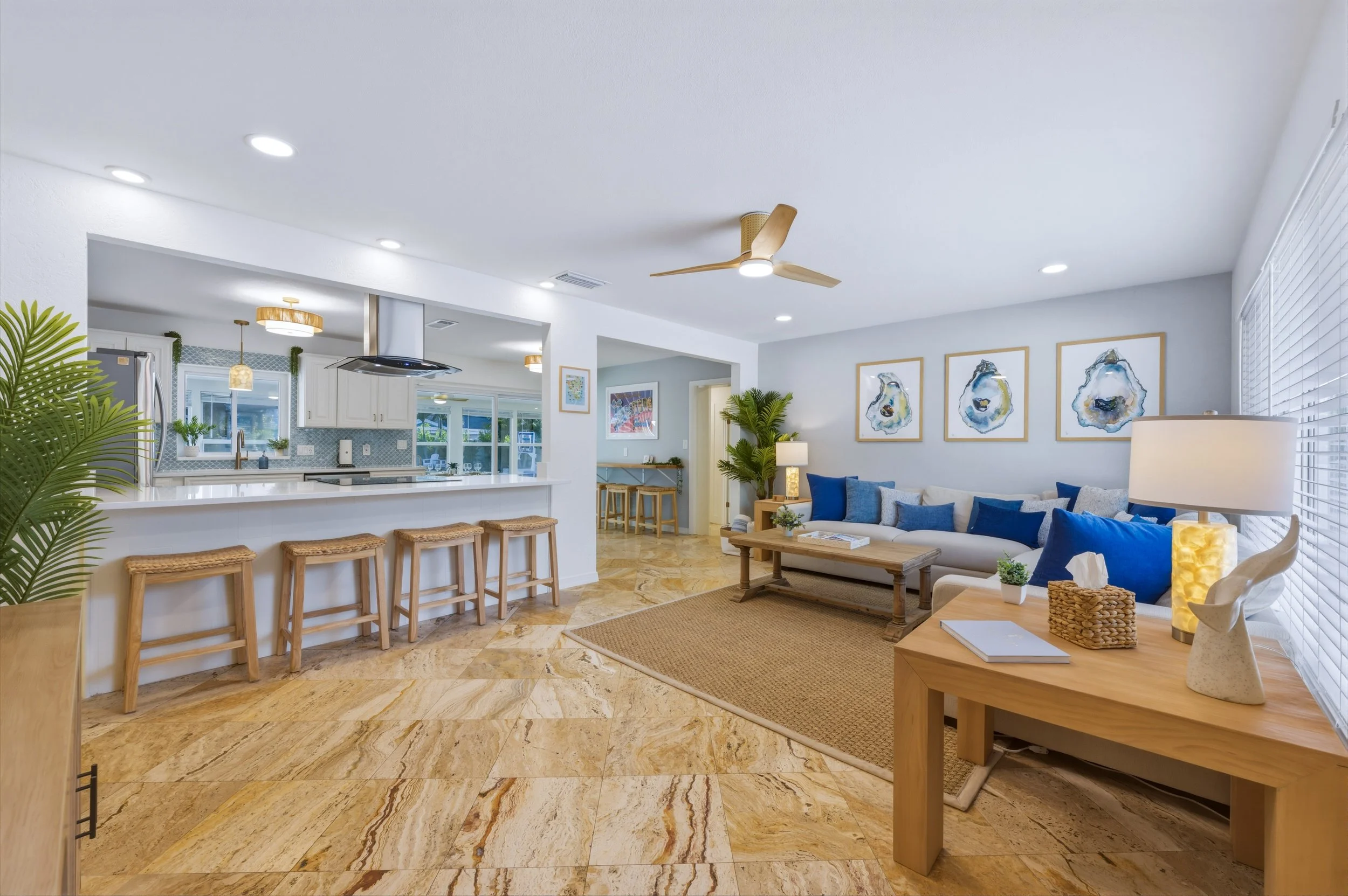 Bright living room with a beige sofa, blue pillows, wooden coffee table, and framed blue abstract art on the white wall. Adjacent to the living room is an open kitchen with white cabinets, a breakfast bar with four wicker stools, and pendant lighting