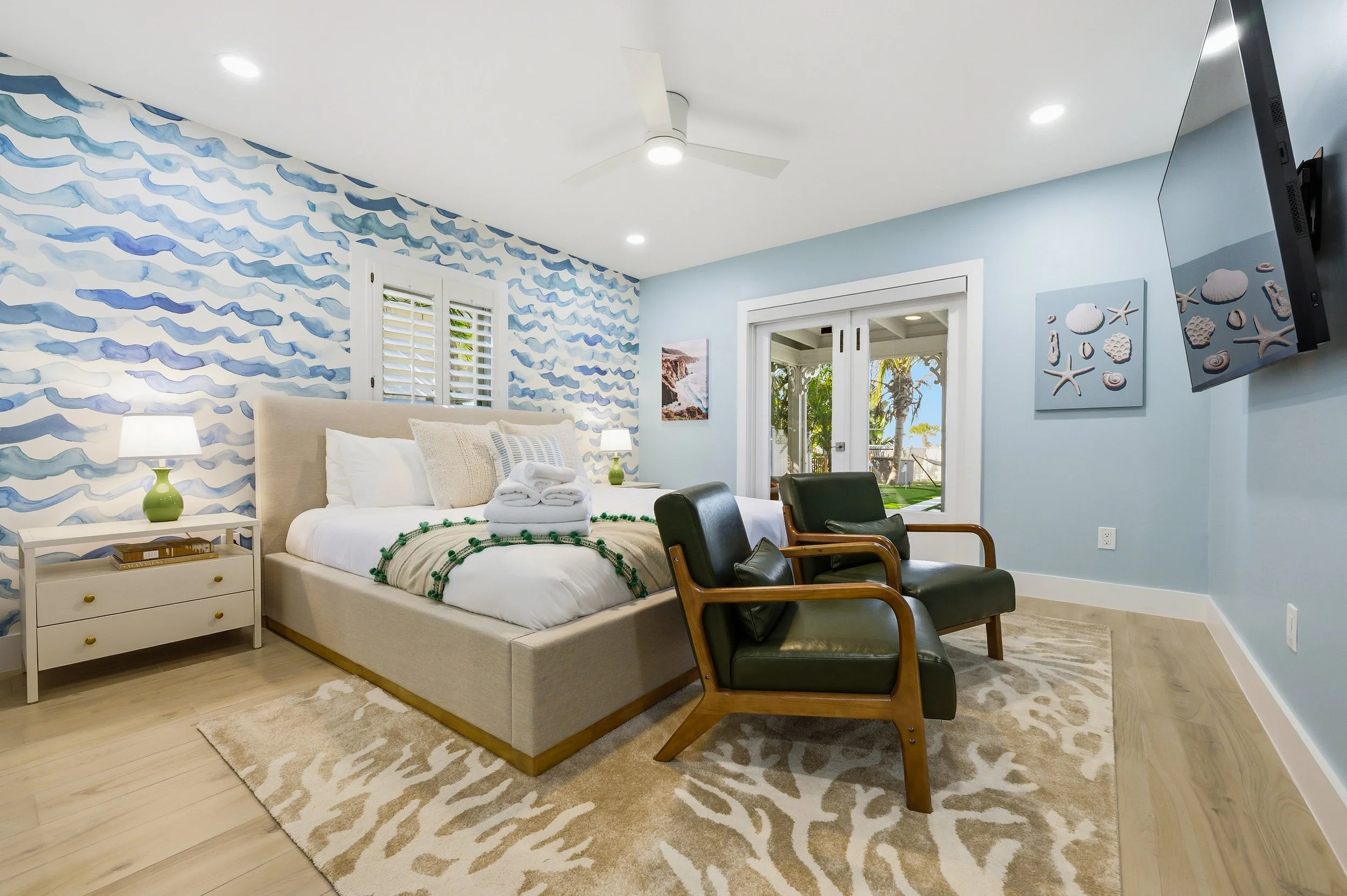 Beach-themed bedroom with a patterned blue wave accent wall, a beige bed with white and beige pillows, towels on the bed, and a view of the outdoors through sliding glass doors showing trees and the ocean.