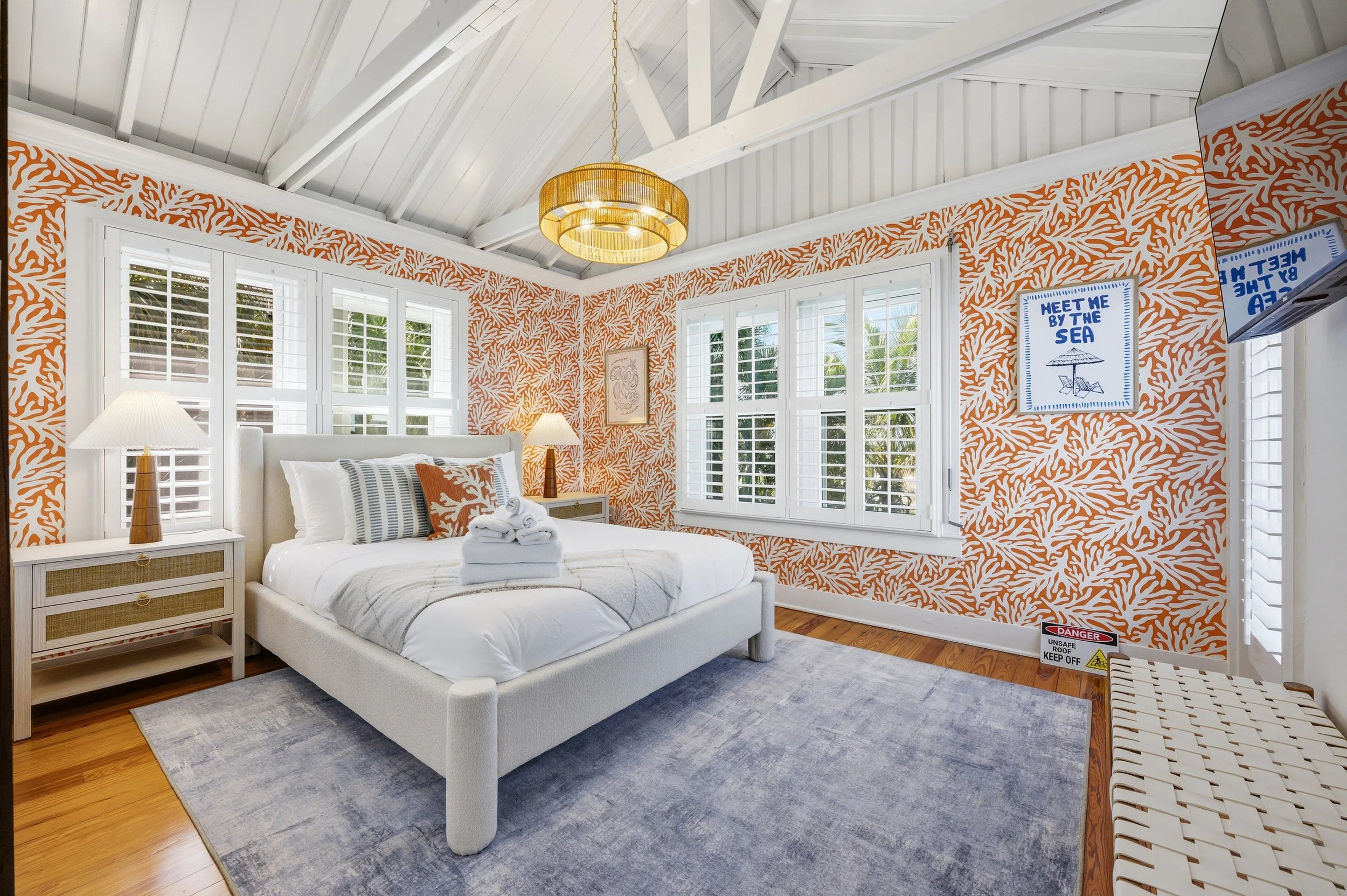 Coastal-inspired bedroom with white bed, orange and white patterned wallpaper, and large windows with white shutters.