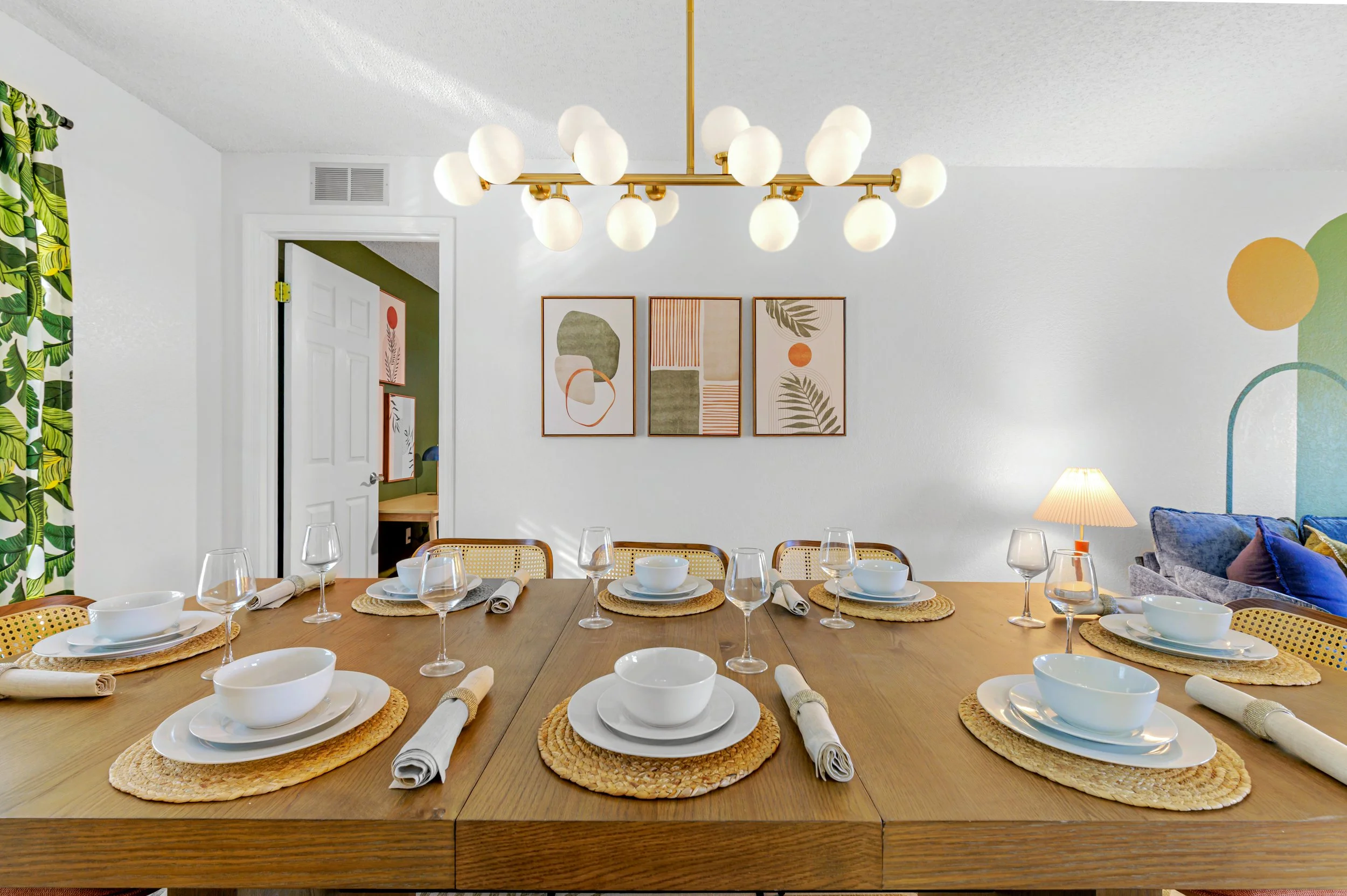 Dining room with a wooden table set with white bowls, plates, glasses, napkins, and woven placemats, decorated with modern artwork on the wall, a gold chandelier, a lamp, and green and white patterned curtains.