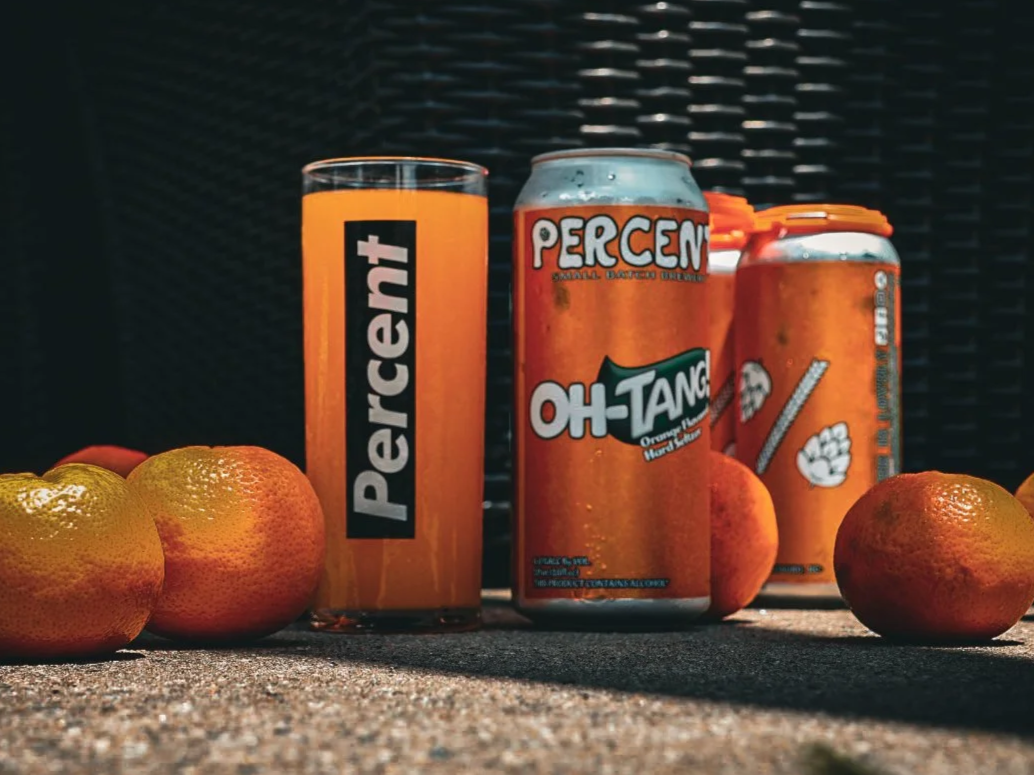 A glass of orange liquid, two cans of orange soda with colorful branding, and several oranges and tangerines are arranged on a textured surface against a dark background.