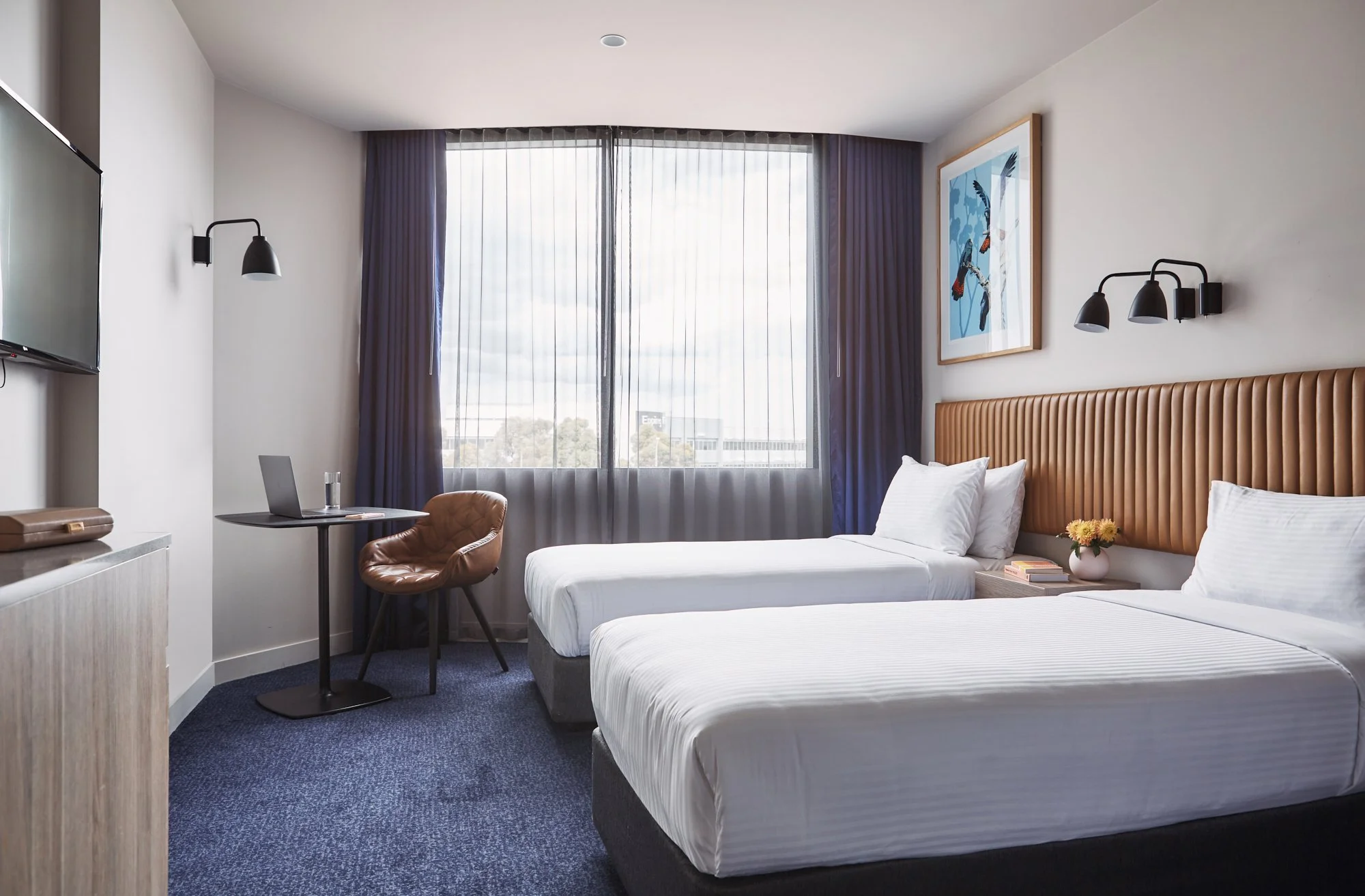 Hotel room with two single beds, white bedding, brown leather chair, small table with laptop, decorative wall art, and large window with sheer curtains.