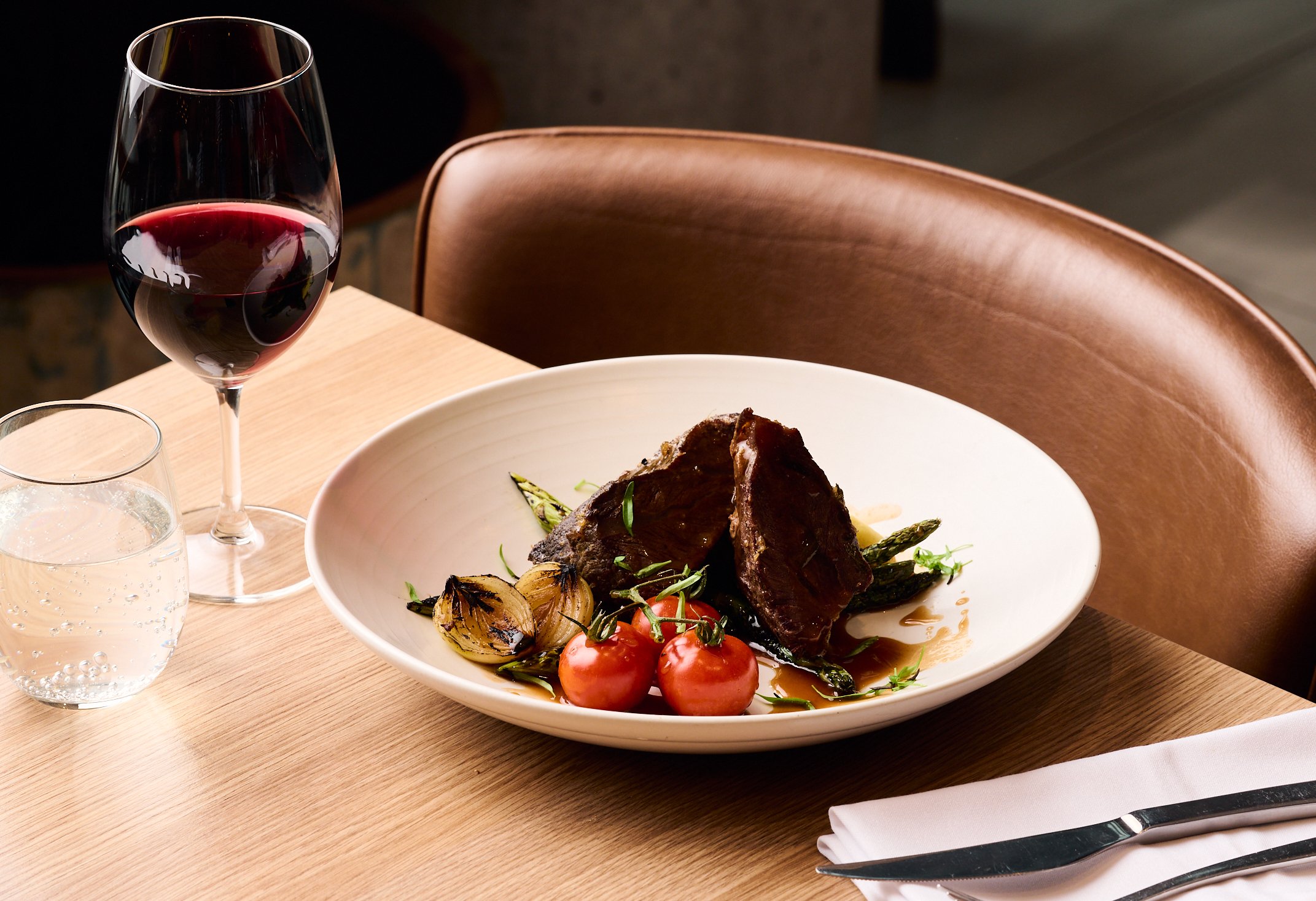 Fine dining table setting with a plate of gourmet beef, vegetables, and a side of wine.