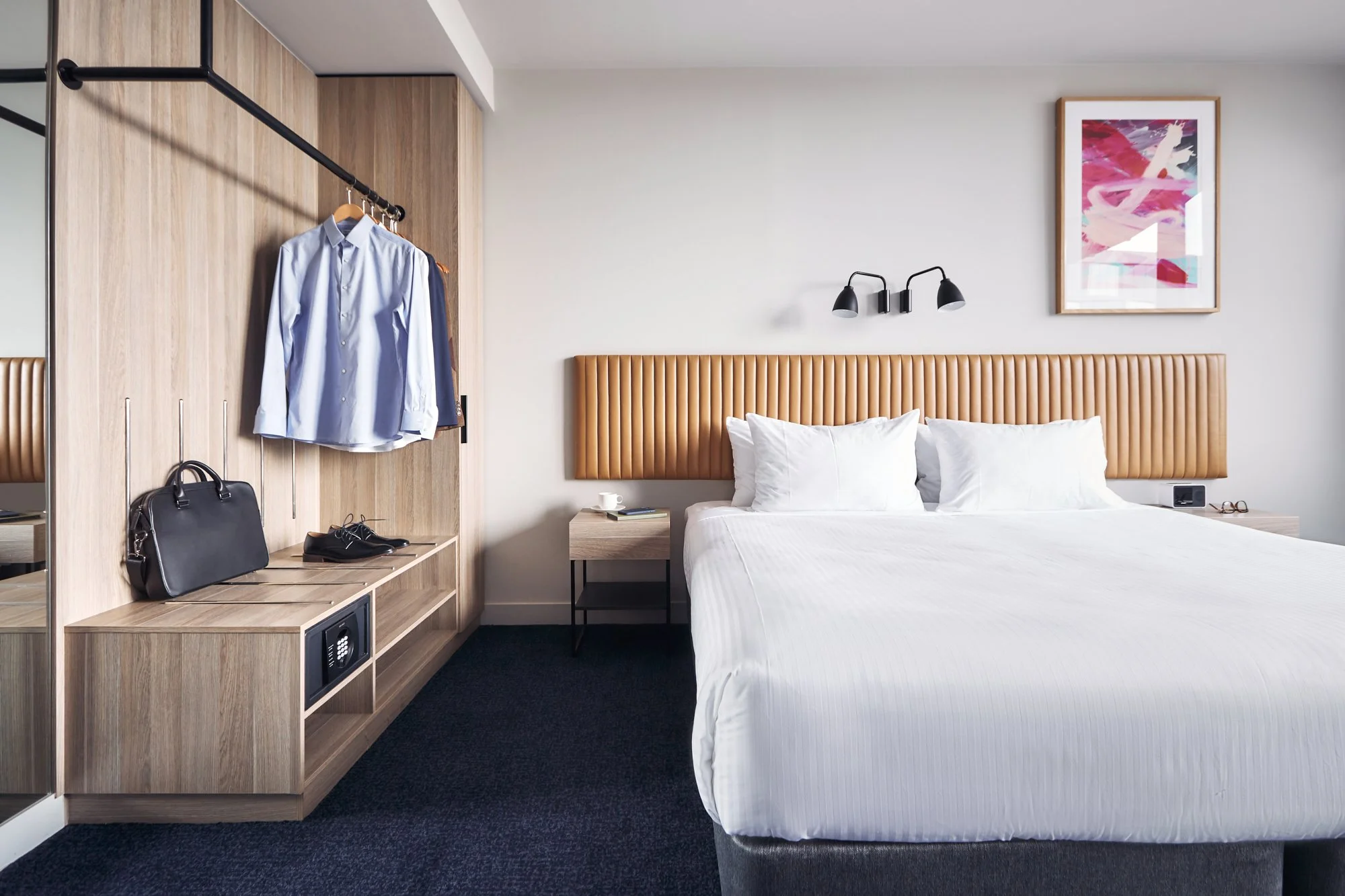 Modern hotel room with a neatly made bed, wooden wardrobe, and bathroom view; includes hanging clothes, a briefcase, and wall art.