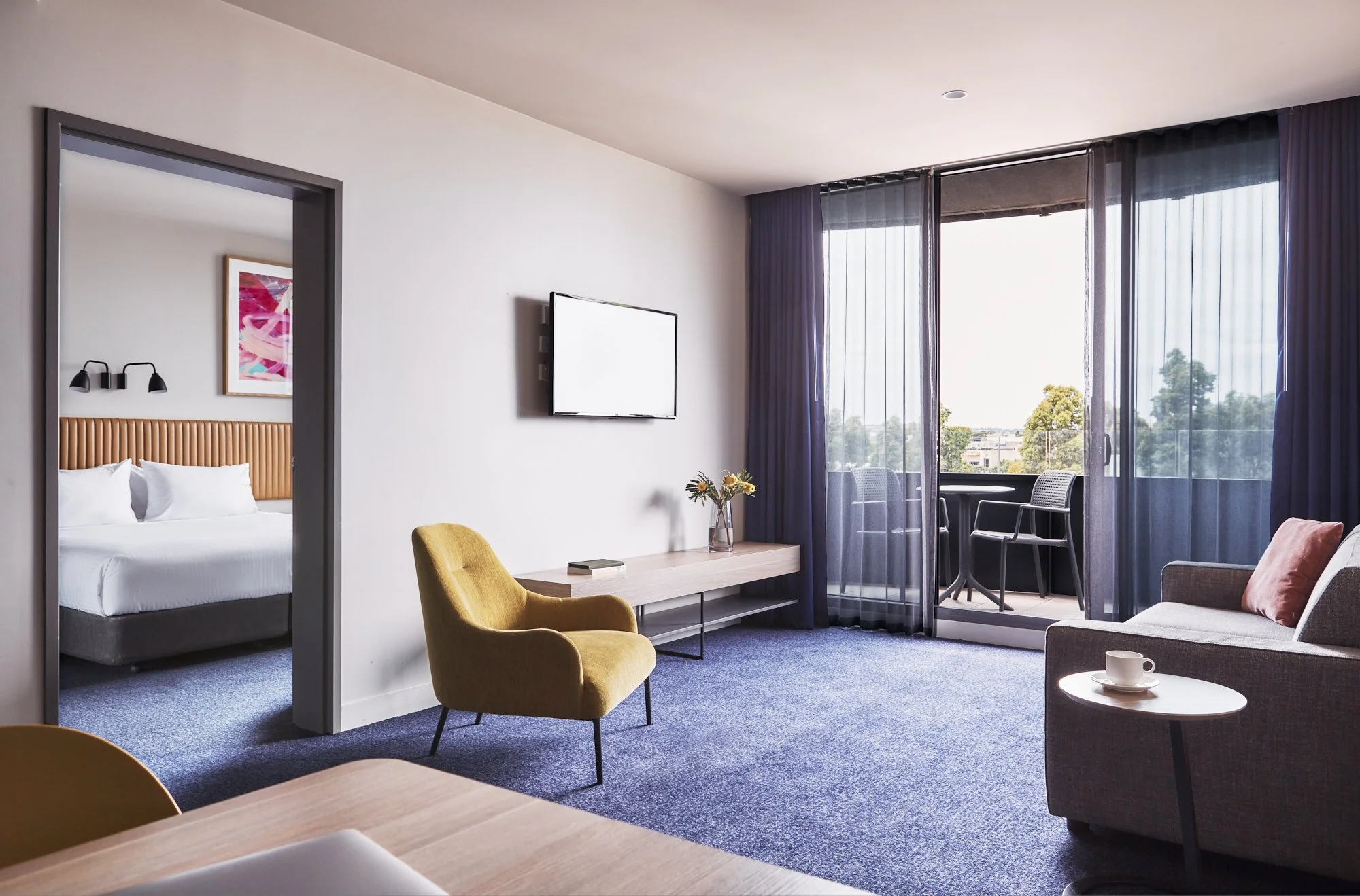 Modern hotel room with a bed in a separate area, a sitting area with a yellow chair, a gray sofa, a wooden table with flowers, a TV, and a balcony with chairs.