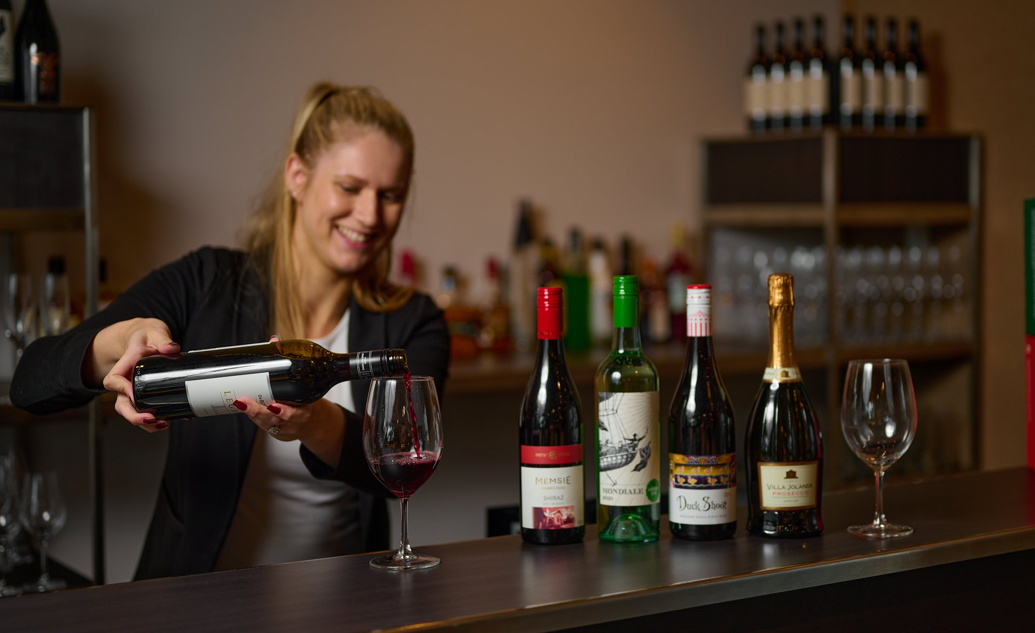 Woman pouring red wine into a glass at a bar, with four bottles of wine lined up on the counter and empty wine glasses nearby.