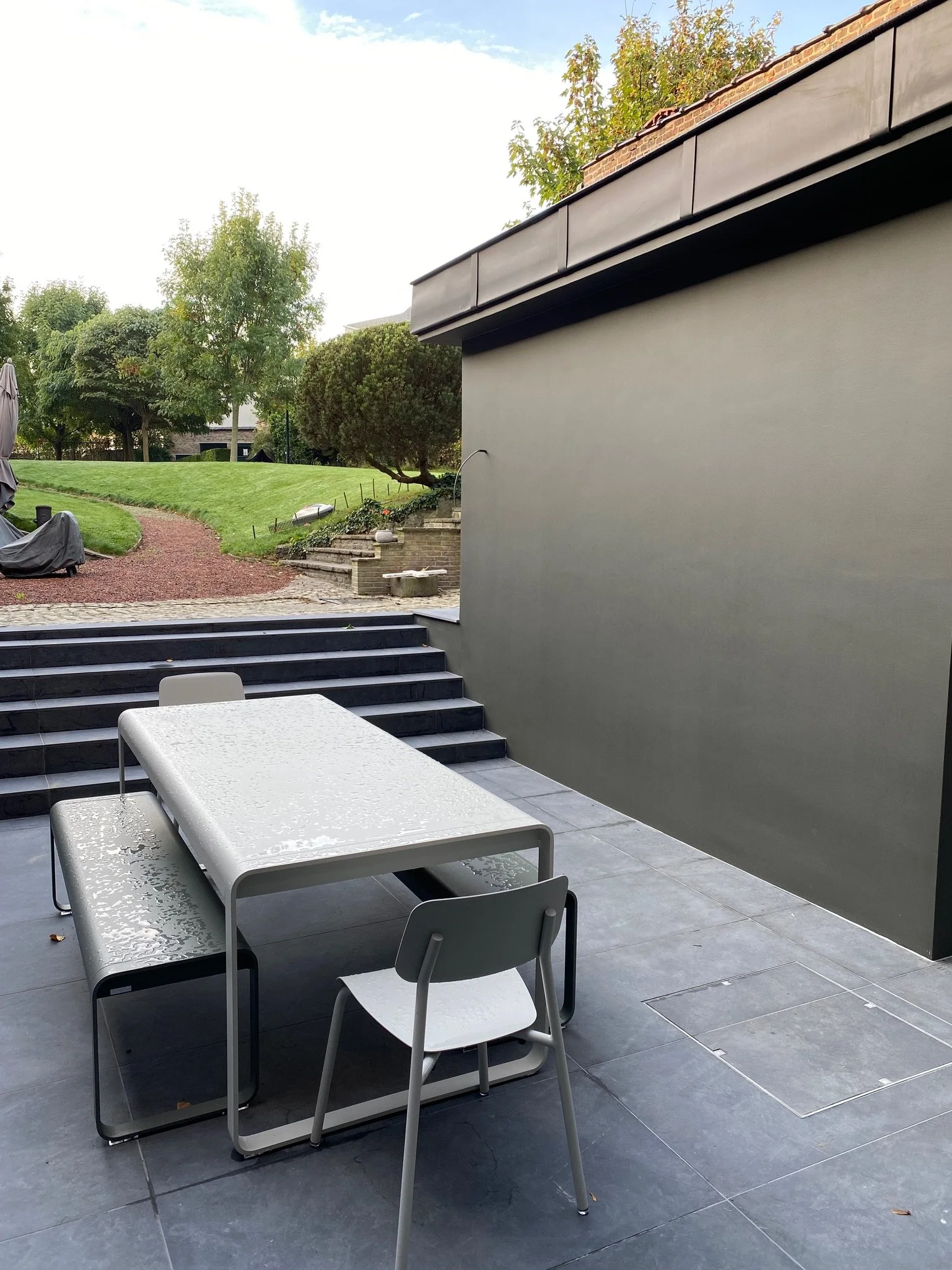 Terrasse avec table et chaises, jardin avec pelouse et arbres à l'arrière-plan.