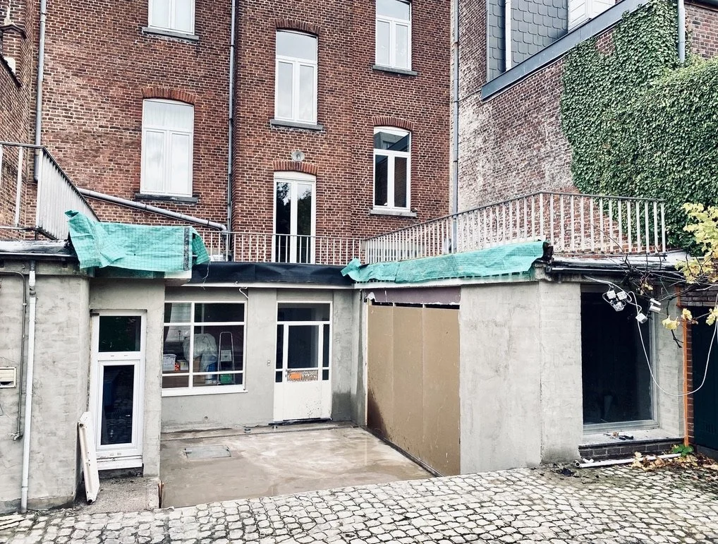 Arrière d'un bâtiment en briques avec fenêtres, terrasse supérieure avec rambarde, toit recouvert de bâches vertes, et un espace pavé en premier plan.