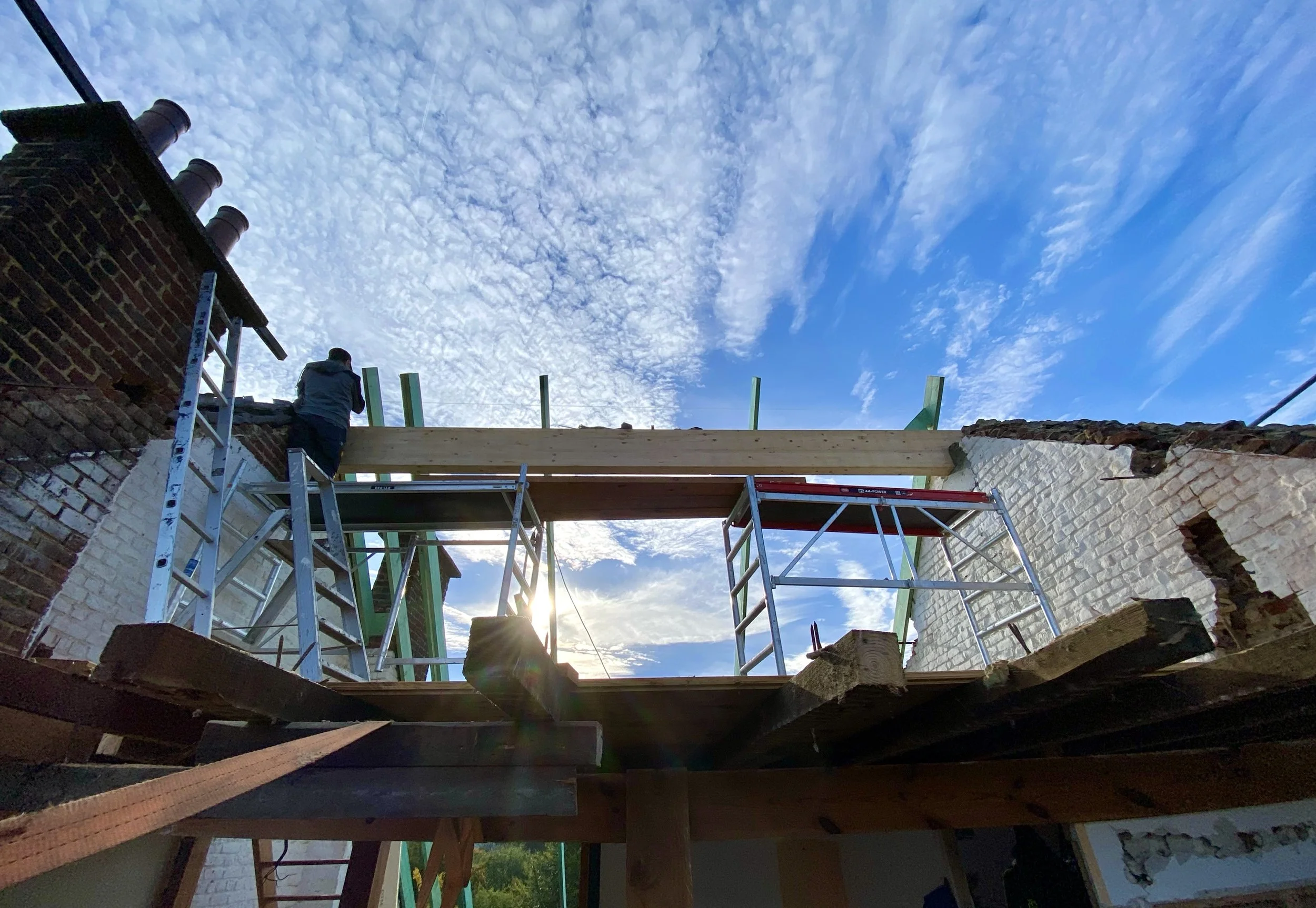 Une scène de construction avec une personne sur une échelle travaillant sur un bâtiment en briques sous un ciel partiellement nuageux.