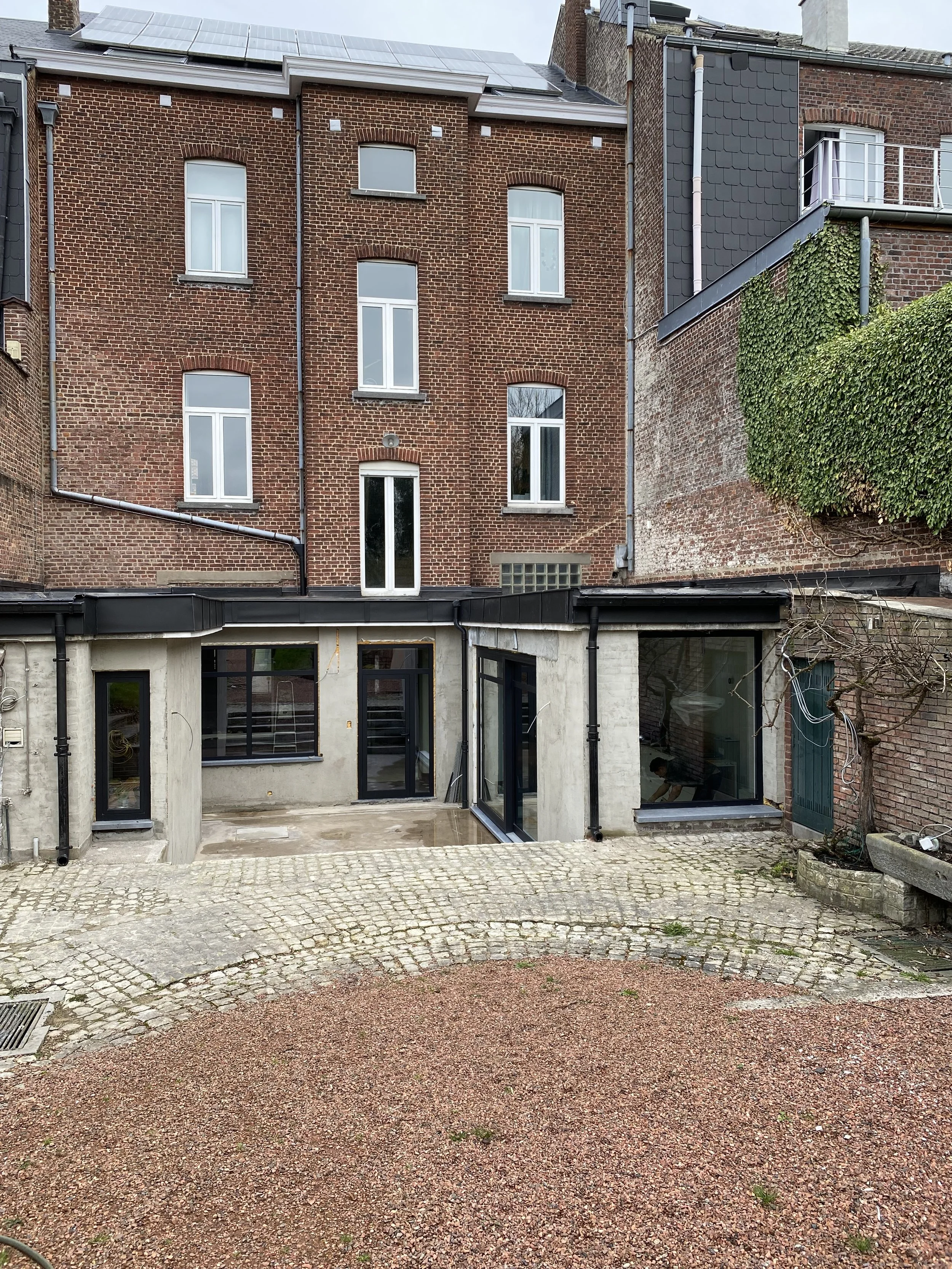 Vue d'une cour arrière avec un bâtiment en briques rouges et des fenêtres modernes. La cour est pavée avec des pierres et un sol en gravier devant le bâtiment.
