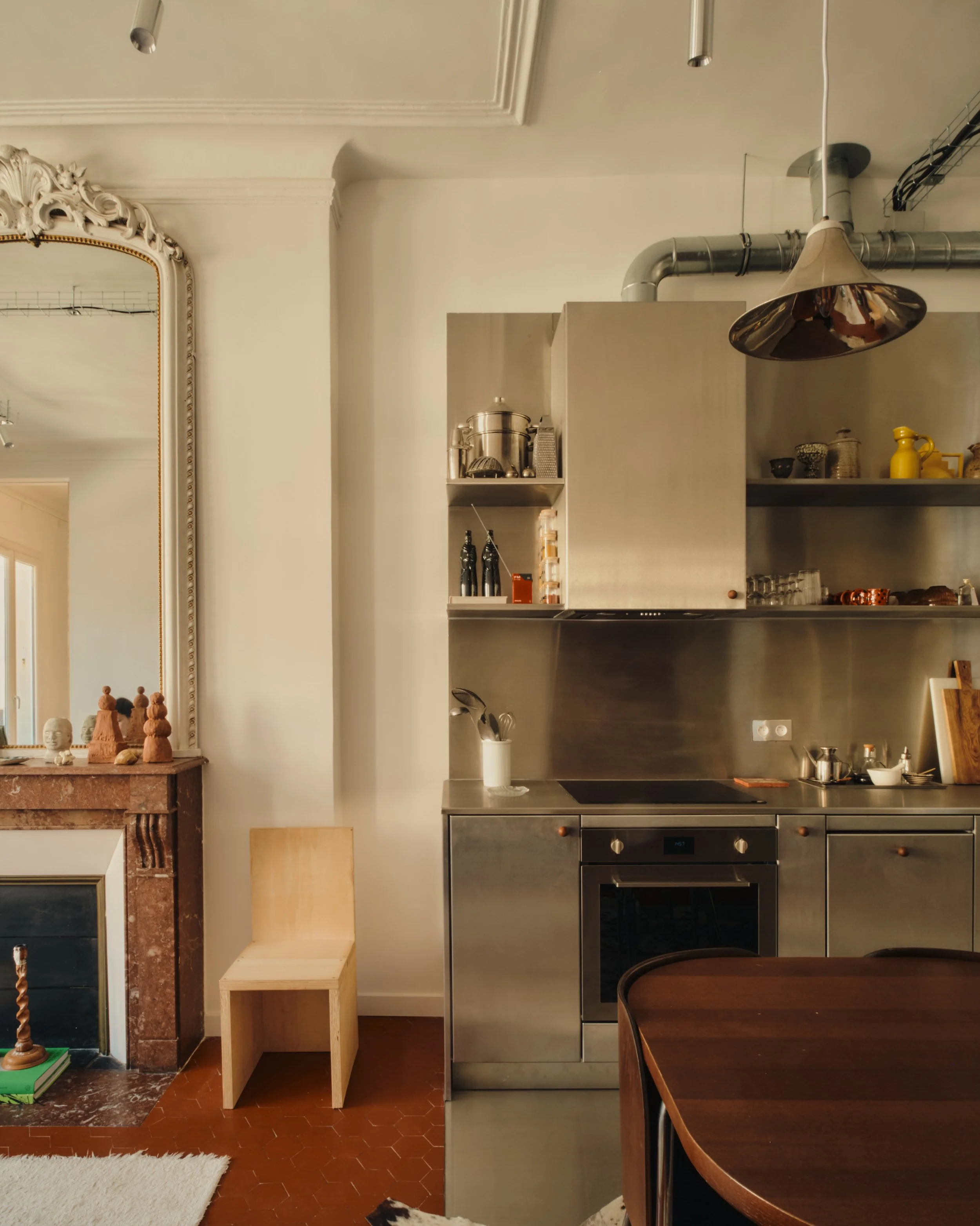 Cuisine moderne en acier inoxydable avec étagères ouvertes, un four intégré, un plan de travail, une table en bois, une cheminée avec miroir au-dessus, et une chaise en bois beige.