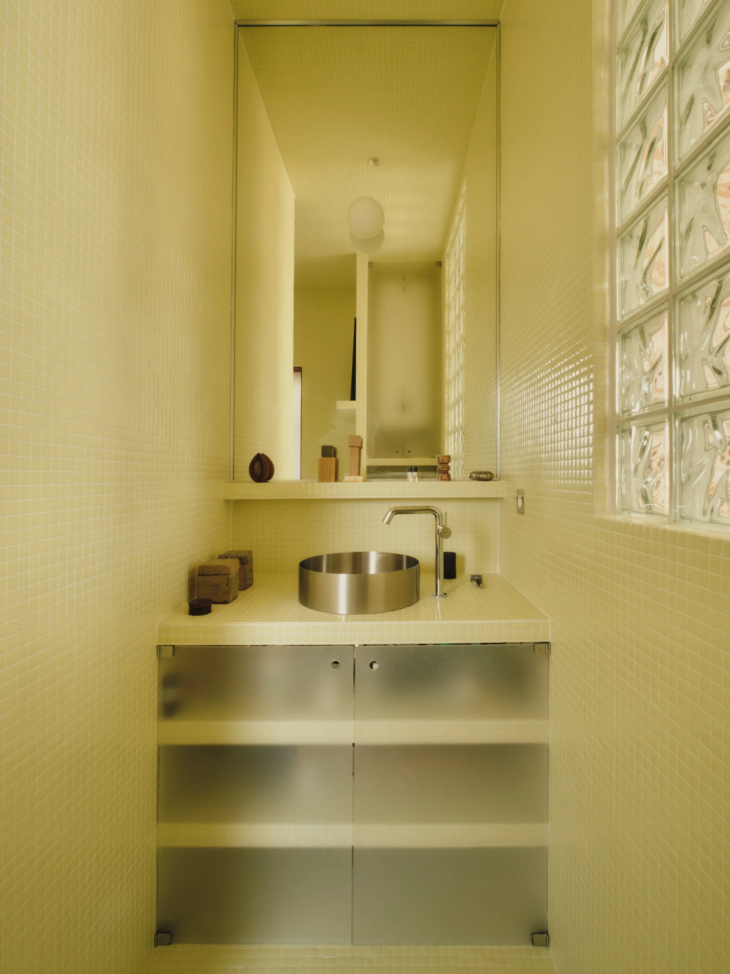 Petite salle de bain avec mursrecouverts de petites carreaux jaunes, lavabo rond en acier inoxydable, étagère en métal au dessous, et un grand miroir rectangulaire au mur. La pièce est éclairée par une fenêtre en blocs de verre.