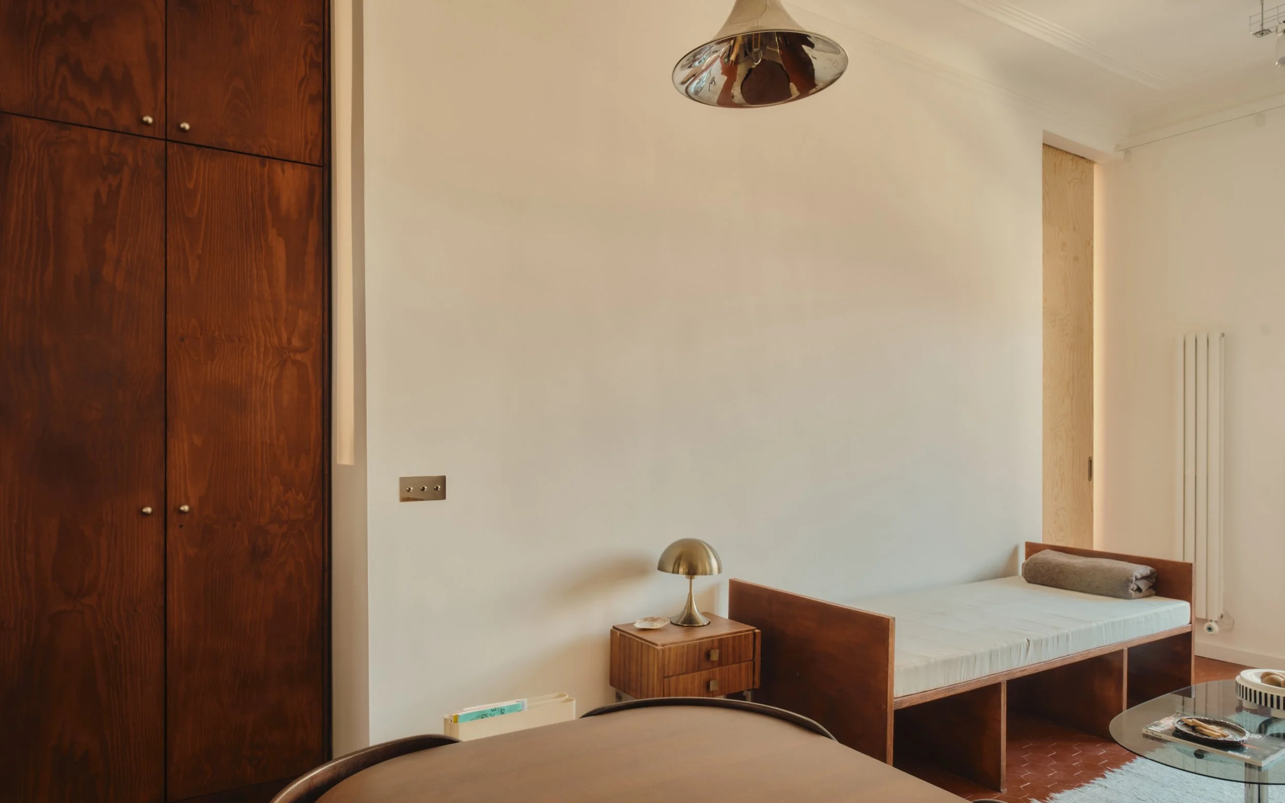 Chambre avec lit simple, table de nuit en bois, lampe en métal, armoire en bois foncé, petite table ronde en verre, décoration minimaliste.