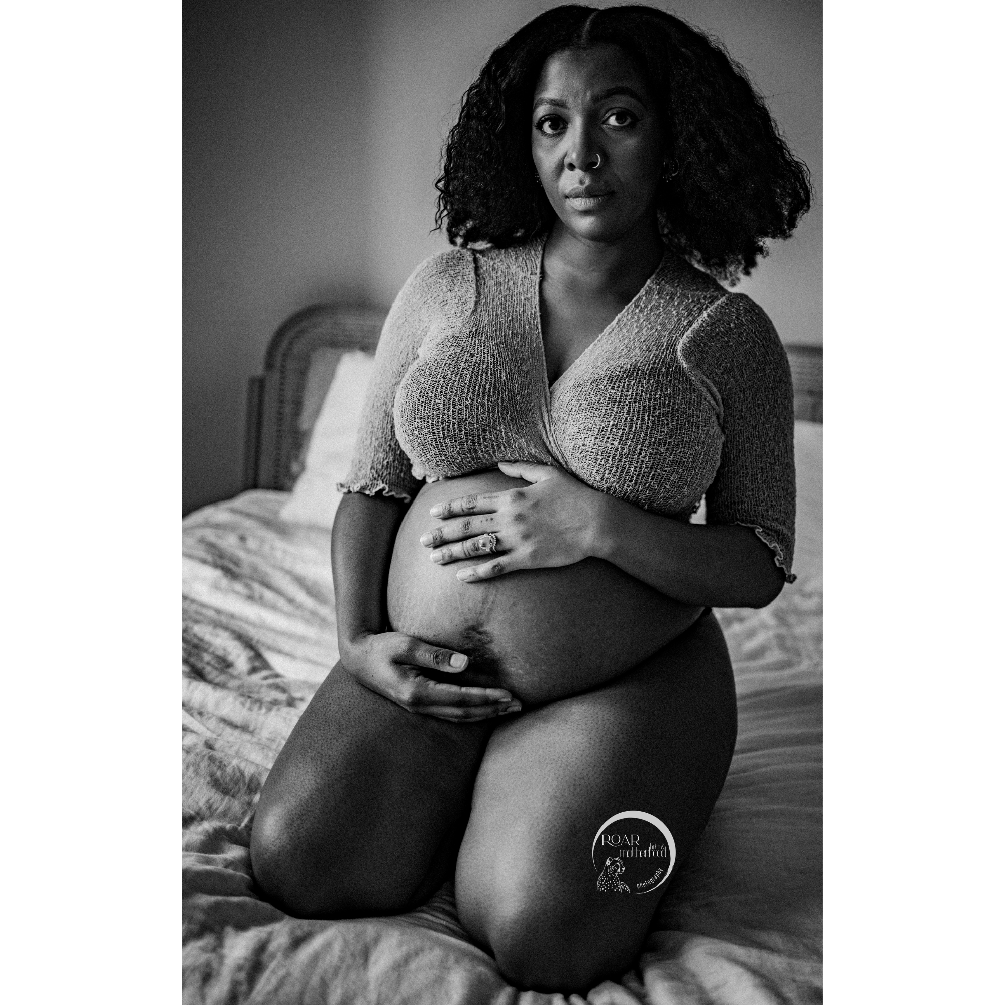 B&W photo of beautiful pregnant black mother, kneeling on the bed, looking into the camera with anxious eyes. She wears knitted cropped wrap top. Phot (c) Roar Birth & Motherhood Photography.
