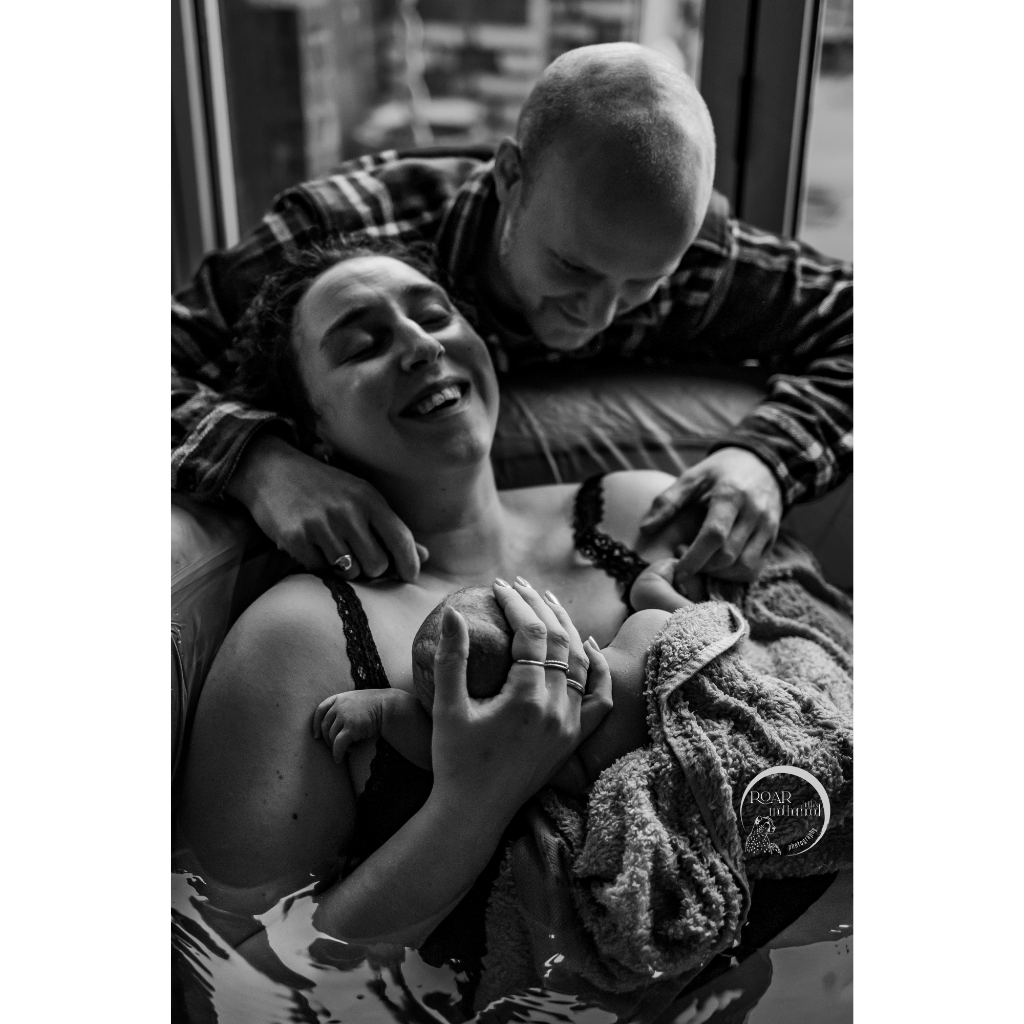 B&W photo of an ecstatic mother who has just birthed her baby in the pool at home. She holds the baby to her chest, joy across her face, while the father gazes at his baby. Photo (c) Roar Birth & Motherhood Photography.