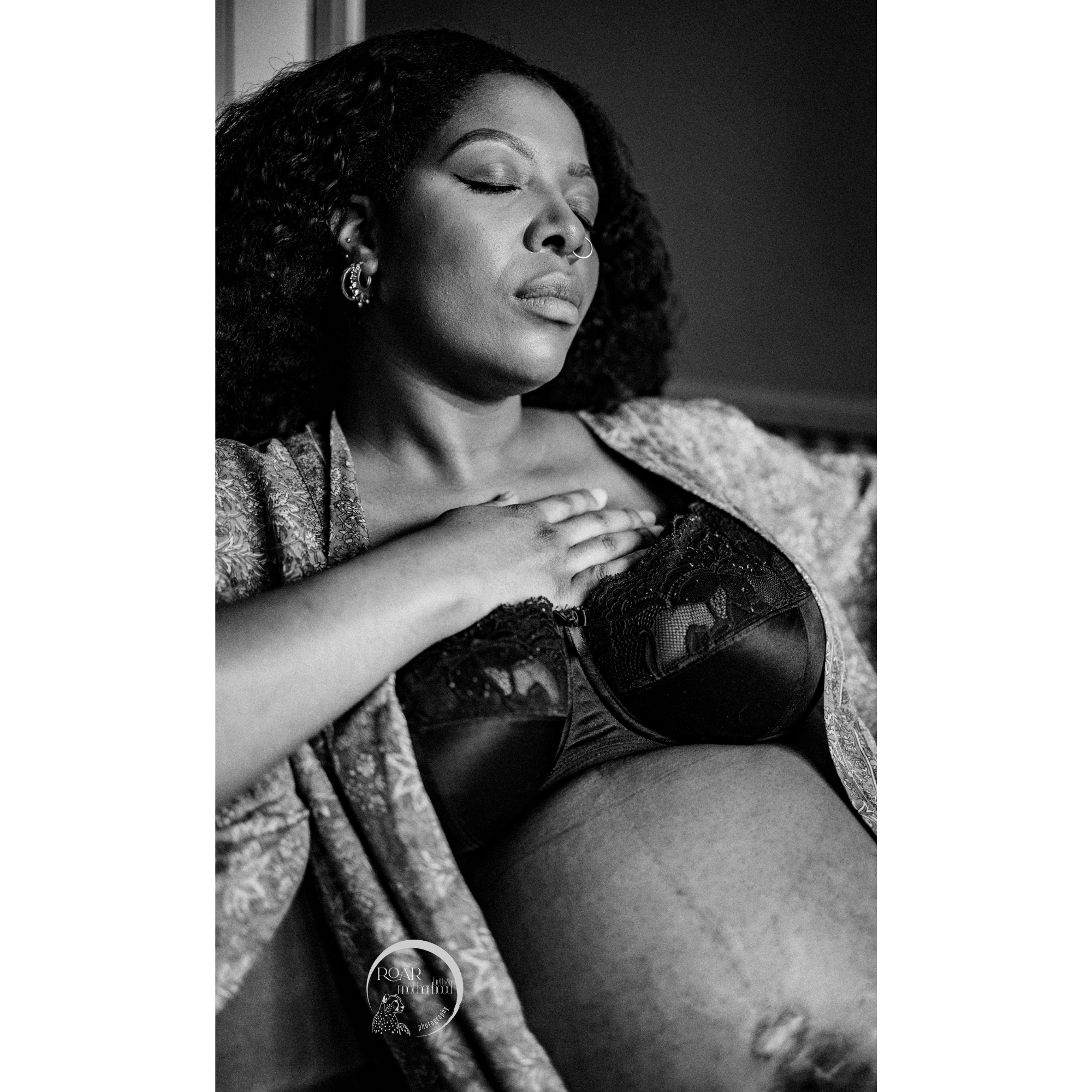 B&W photo of pregnant black mother, eyes closed, hand on her heart, deep in thought. She wears black bra and loose robe and has ears and nose piercings. Photo (c) Roar Birth & Motherhood Photography.