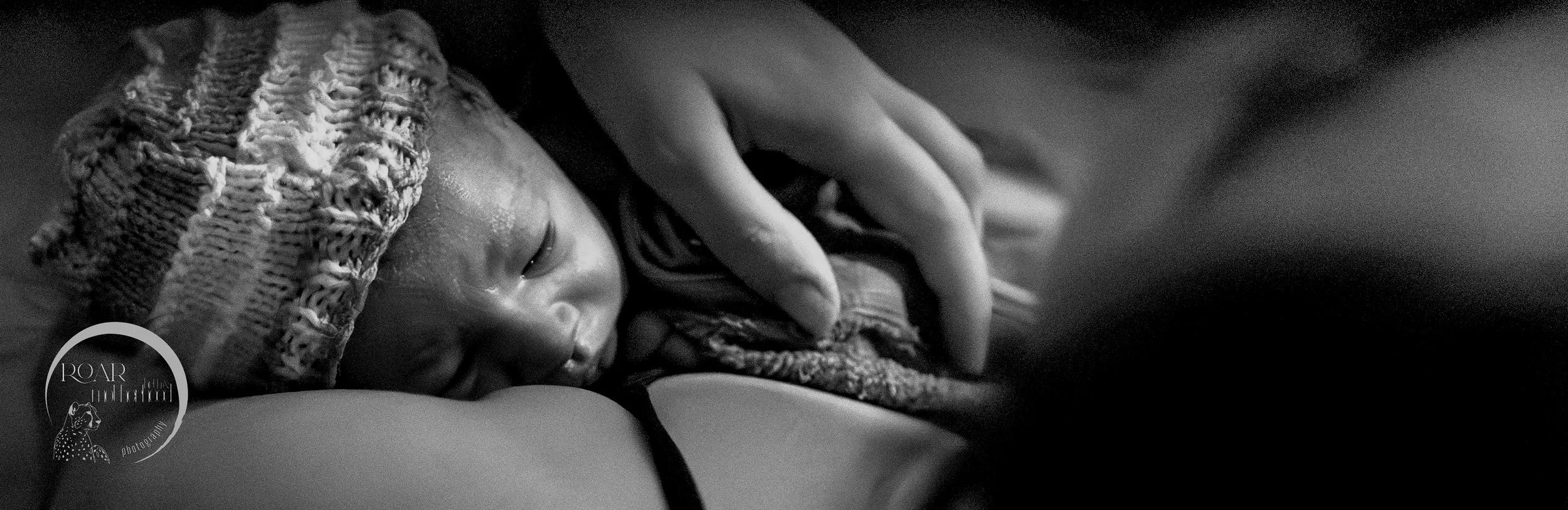 B&W photo of newborn baby being held close by his mother, having just been born in the pool at home. The baby wears a knitted hat and is calm, while the mother rests her head back as she takes stock. Photograph (c) Roar Birth & Motherhood Photography