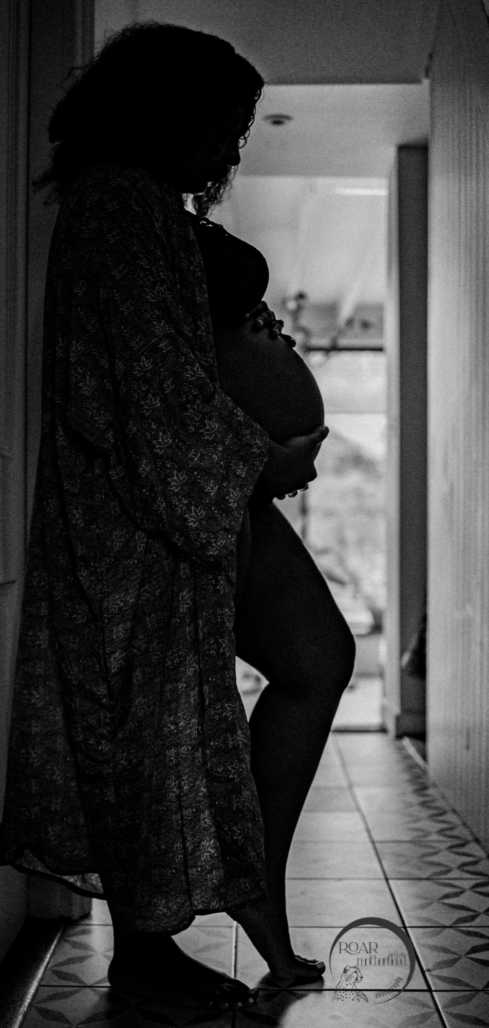 Black and white photo of glorious pregnant mother in silhouette in hallway. She wears a loose gown and cradles her bump while deep in thought.