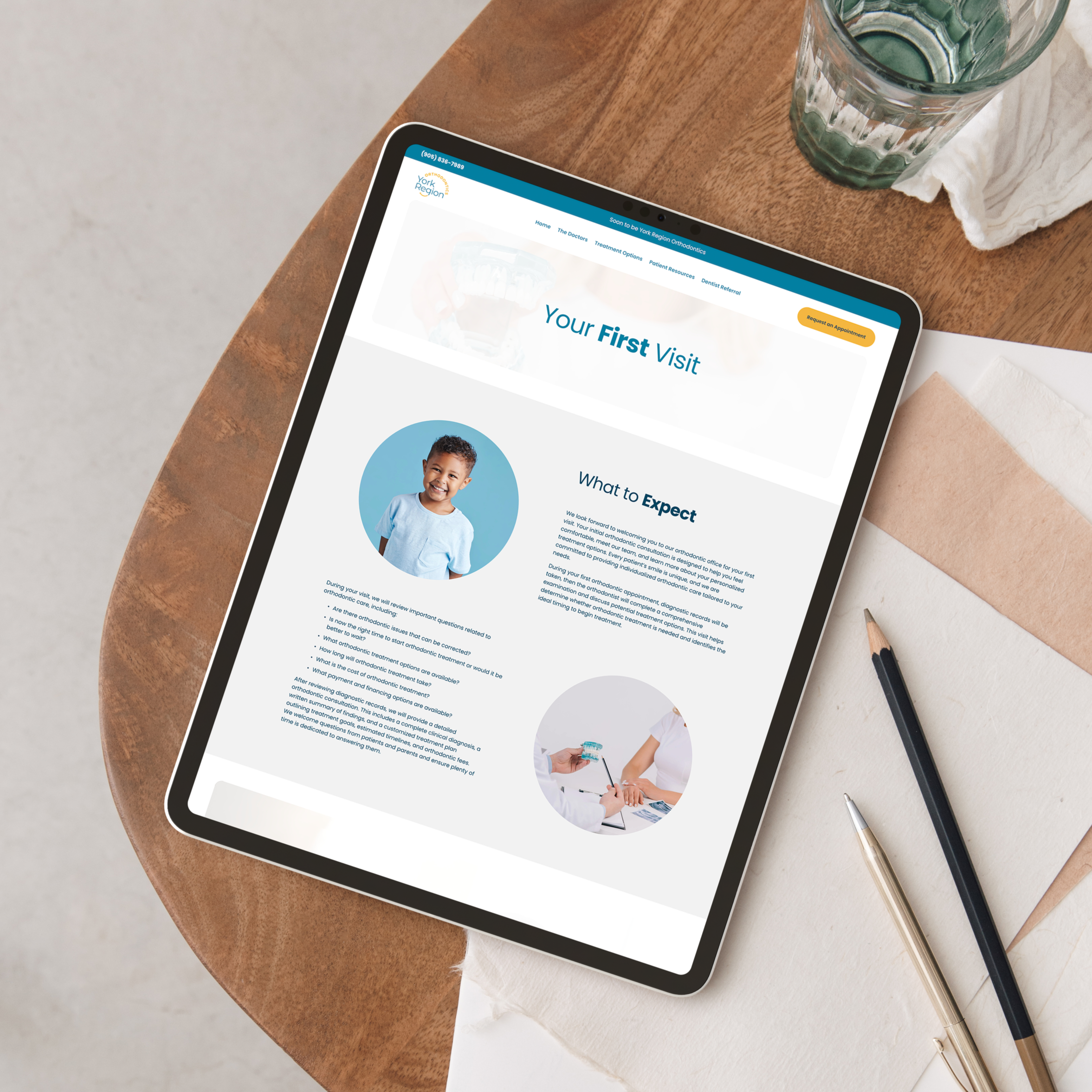 Tablet displaying a dental clinic's webpage titled "Your First Visit" on a wooden table with a glass of water, a pen, and some papers nearby.
