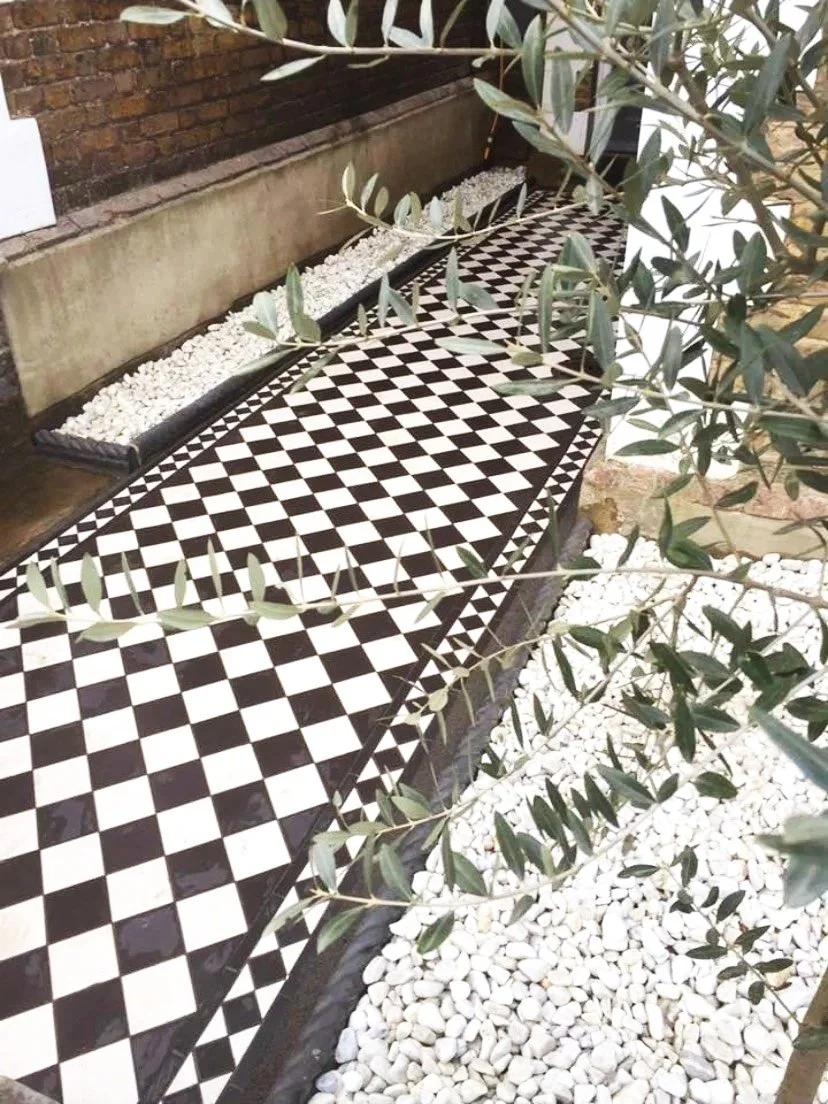 A black and white checkered Victorian tile pathway with white pebble border on either side, bordered by a brick wall and greenery with leaves.