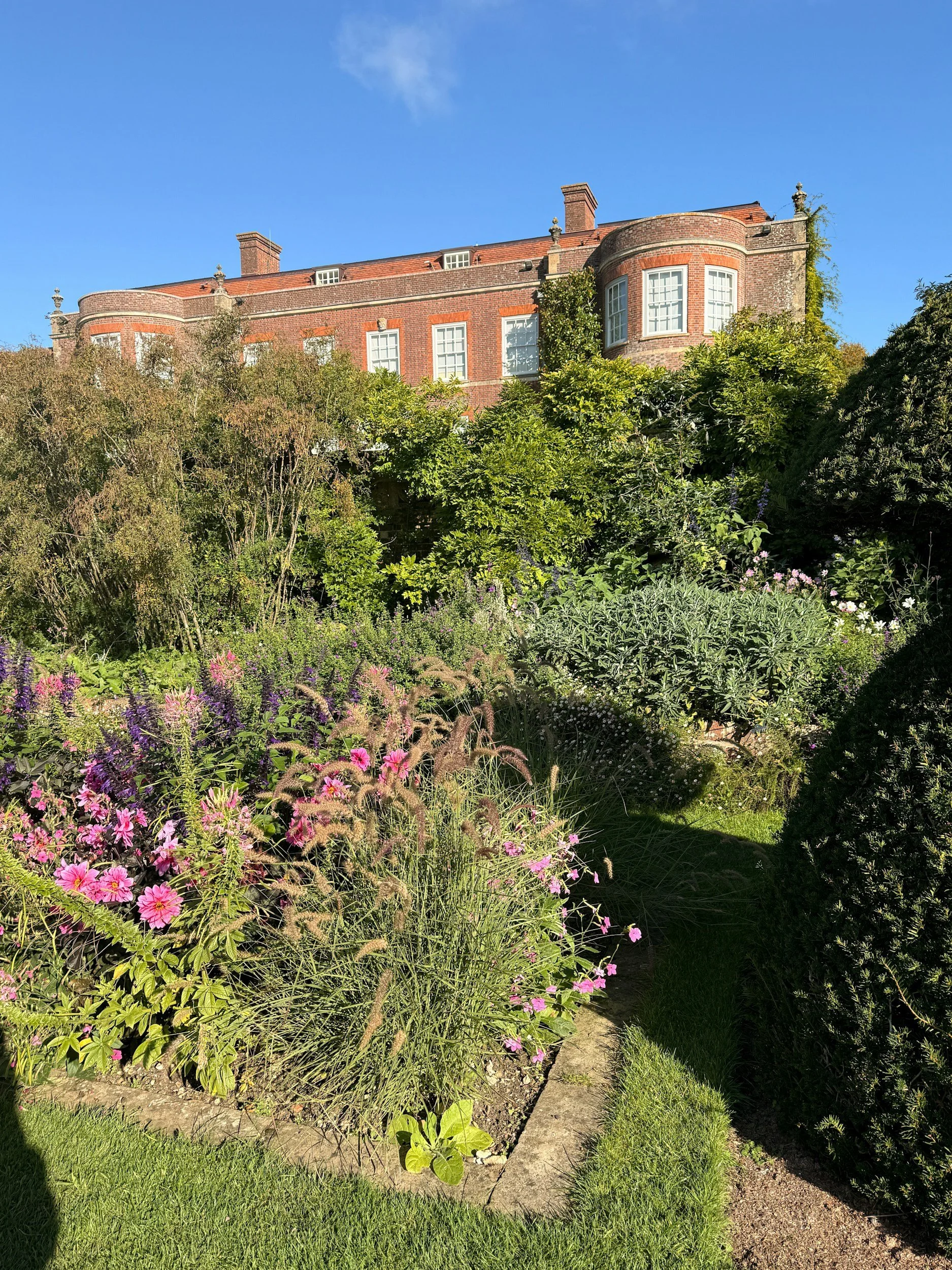 A large brick house with multiple windows, surrounded by lush green trees and a colorful flower garden in the foreground, under a bright blue sky.