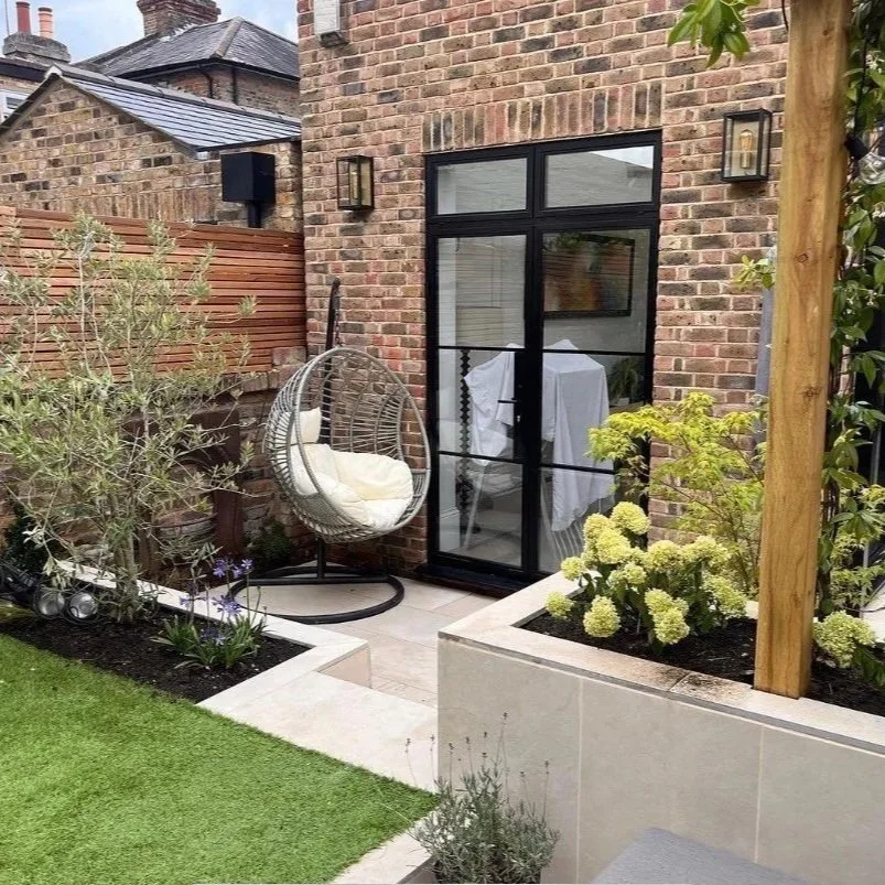 The Berkshire Garden & Design Co. Ltd garden landscape and hardscape design in Windsor, UK.A small outdoor patio area with a modern black metal-framed glass door, a hanging wicker chair with a cushion, surrounded by garden plants.
