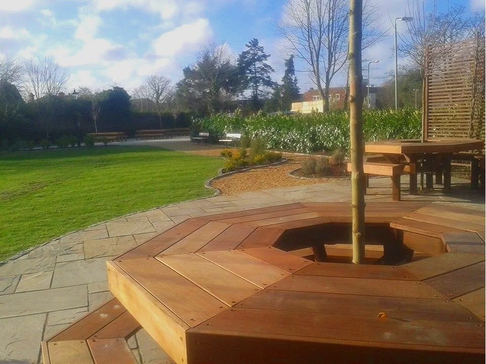 A park with a central tree surrounded by a wooden bench, green grass, and a sidewalk, with benches and trees in the background under a cloudy sky.