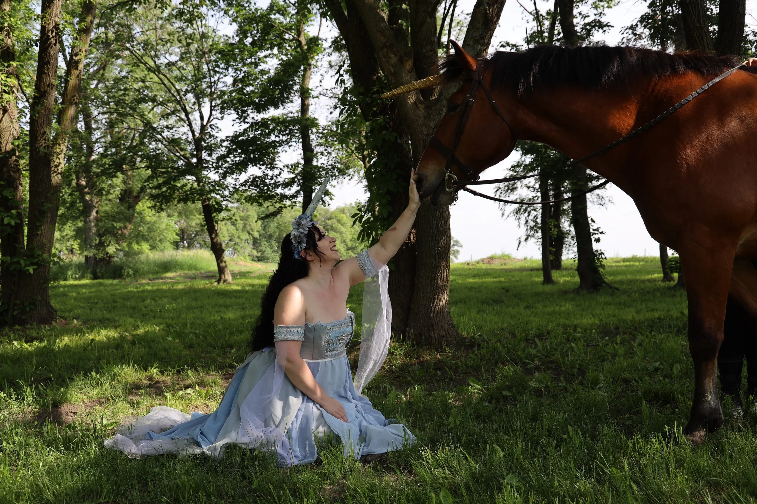 A woman dressed in a blue and gray historical dress kneels on the grass and touches a brown horse's face while sitting outdoors in a park with trees.