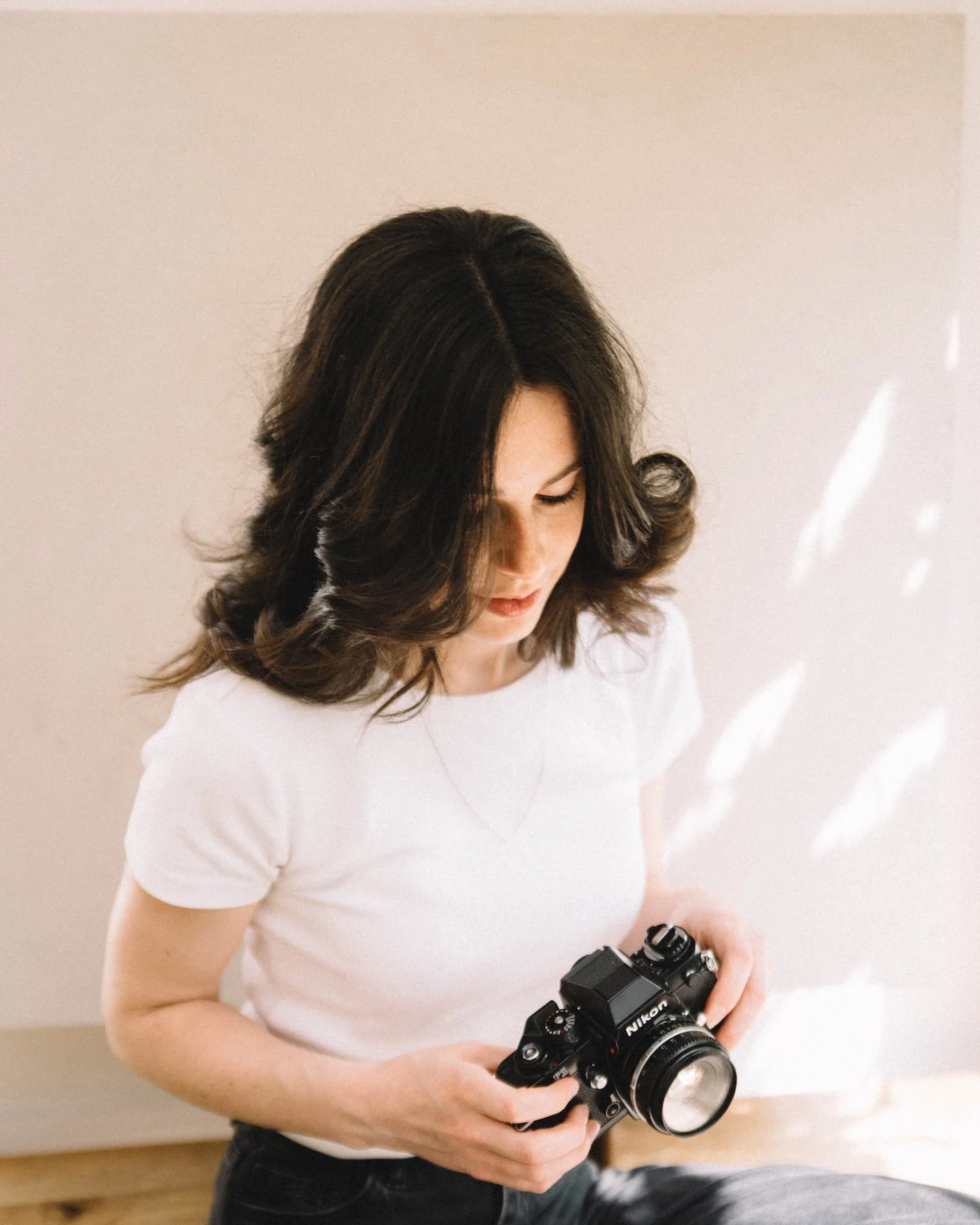 Woman with shoulder-length dark hair wearing a white t-shirt looking down at a Nikon camera in her hands indoors.