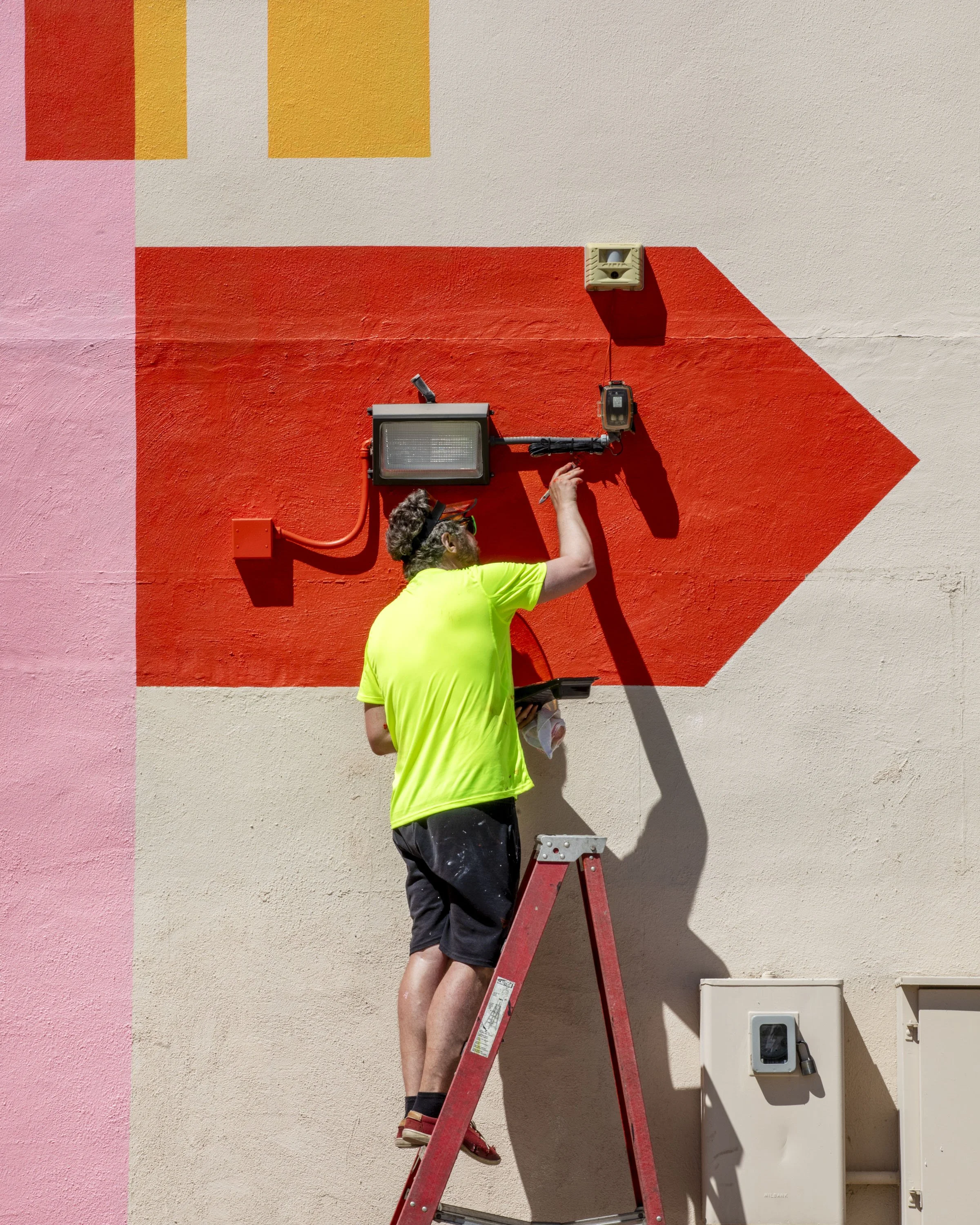 A man on a ladder painting a red arrow on a wall, with other electrical equipment attached above