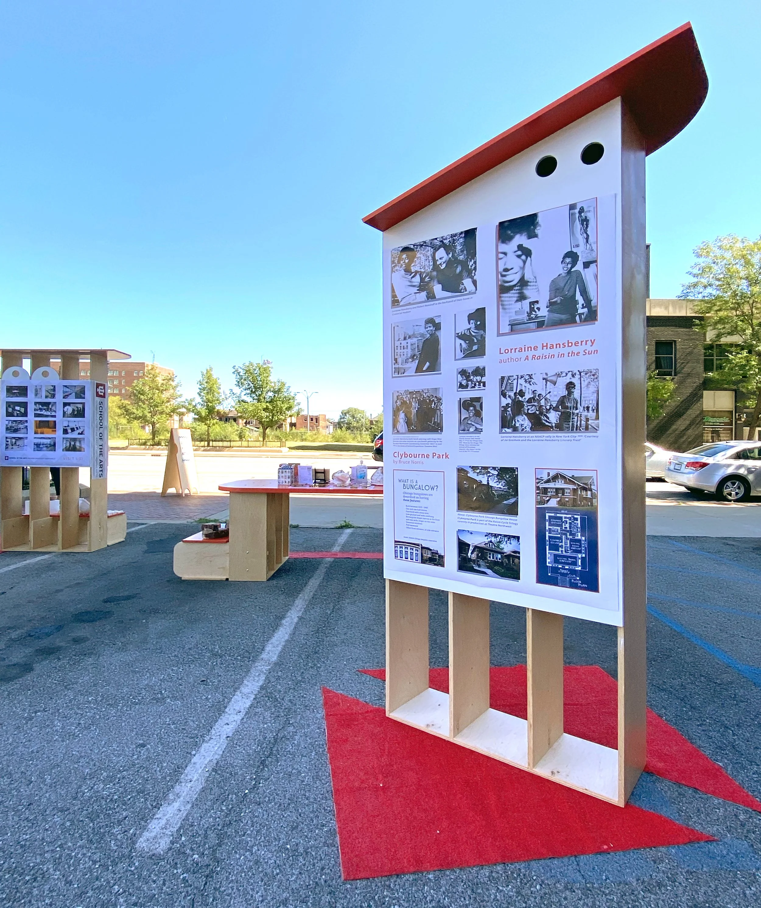 Outdoor art exhibit with display boards featuring photographs and information, set on a pavement with a red carpet and wooden display structures under a clear blue sky.