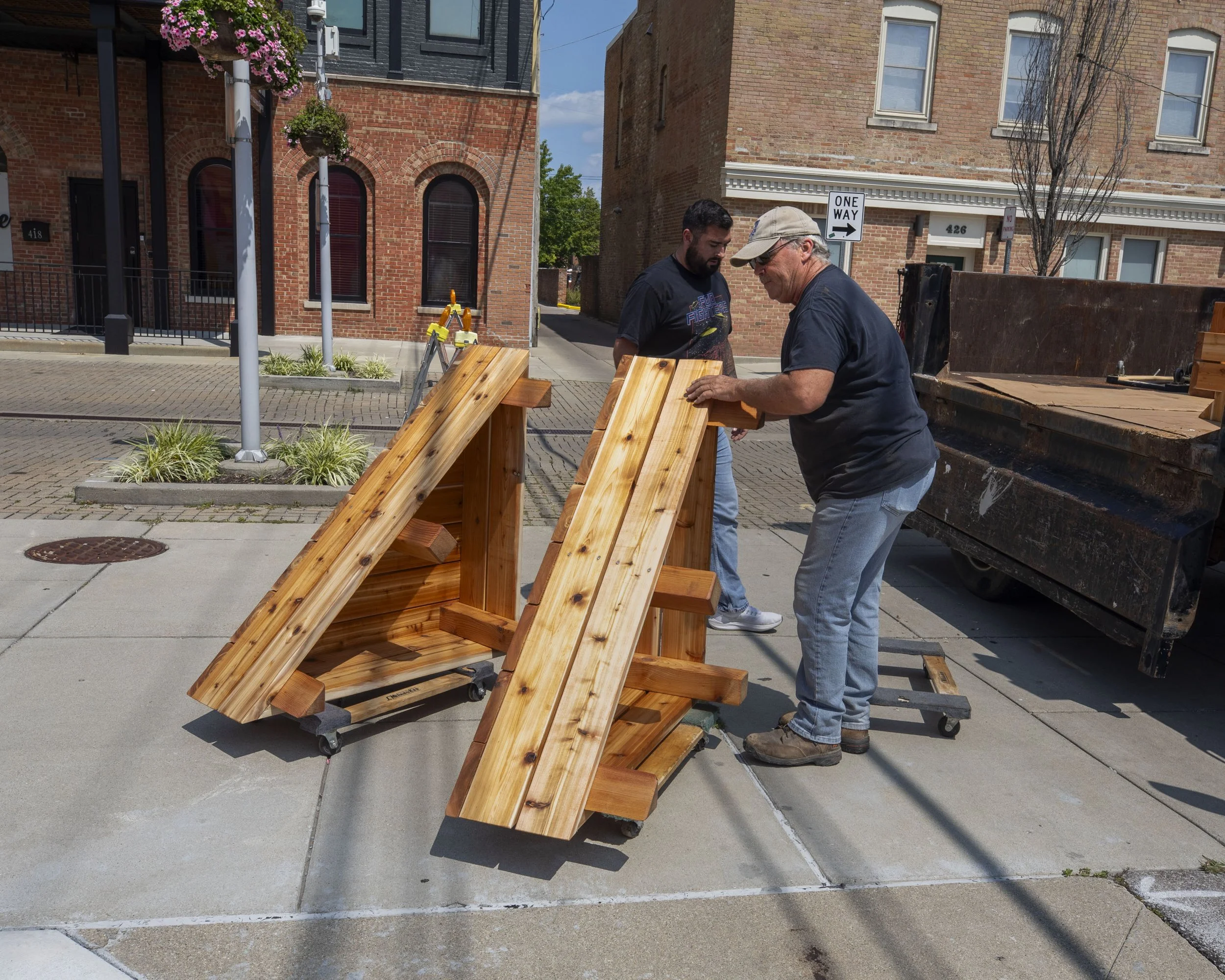 Two men are assembling wooden tables on a sidewalk in an urban area with brick buildings in the background. One man is wearing a cap and glasses, while the other has dark hair and a beard.