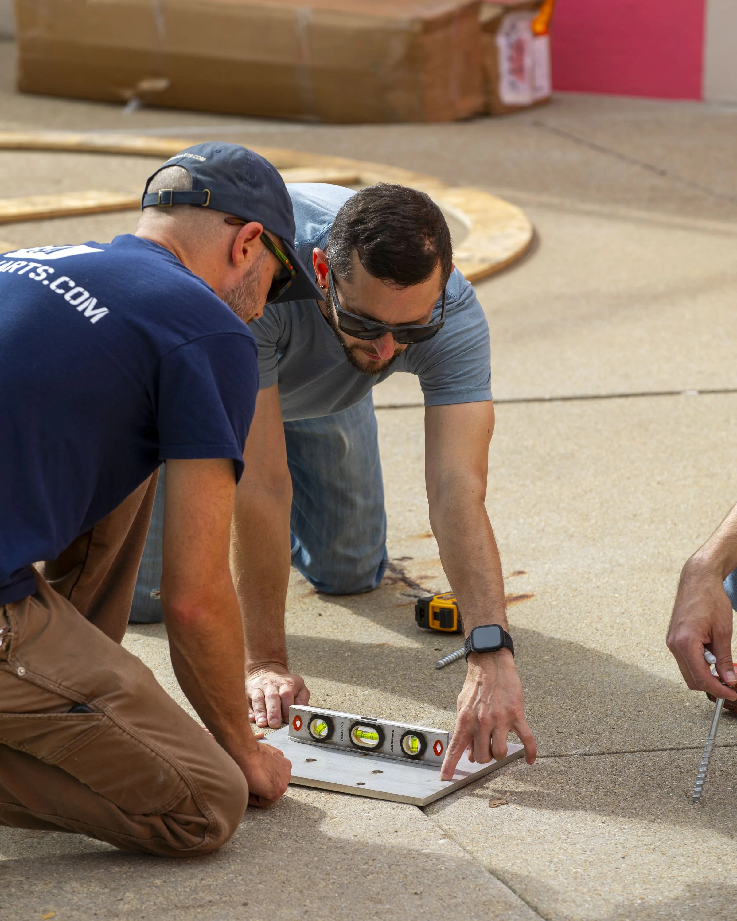 Two men working on a construction project outdoors. One man is counting on his fingers, the other is pointing at a level tool on the ground. Various tools are nearby.