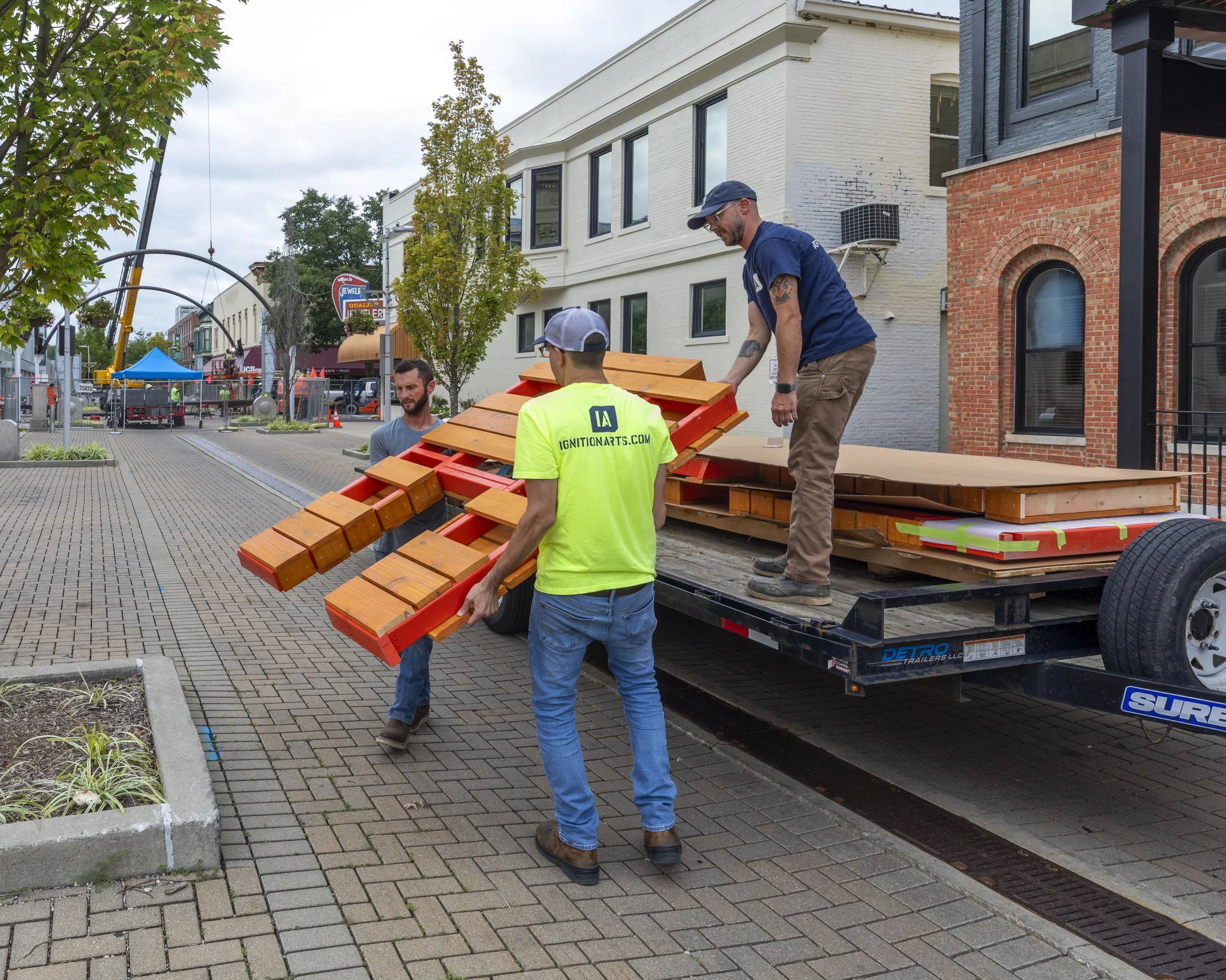 Three men unload wooden benches from a flatbed trailer on a city sidewalk. One man is standing on the trailer, another is handing objects to a man in a bright yellow shirt, and a third man stands on the sidewalk. The background shows trees, buildings, and street tents.