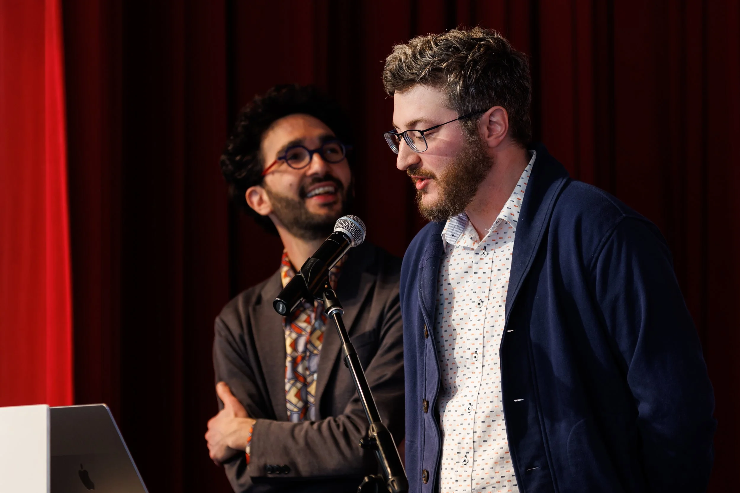 Two men engaged in conversation at a public speaking event, with one speaking into a microphone and the other listening attentively.