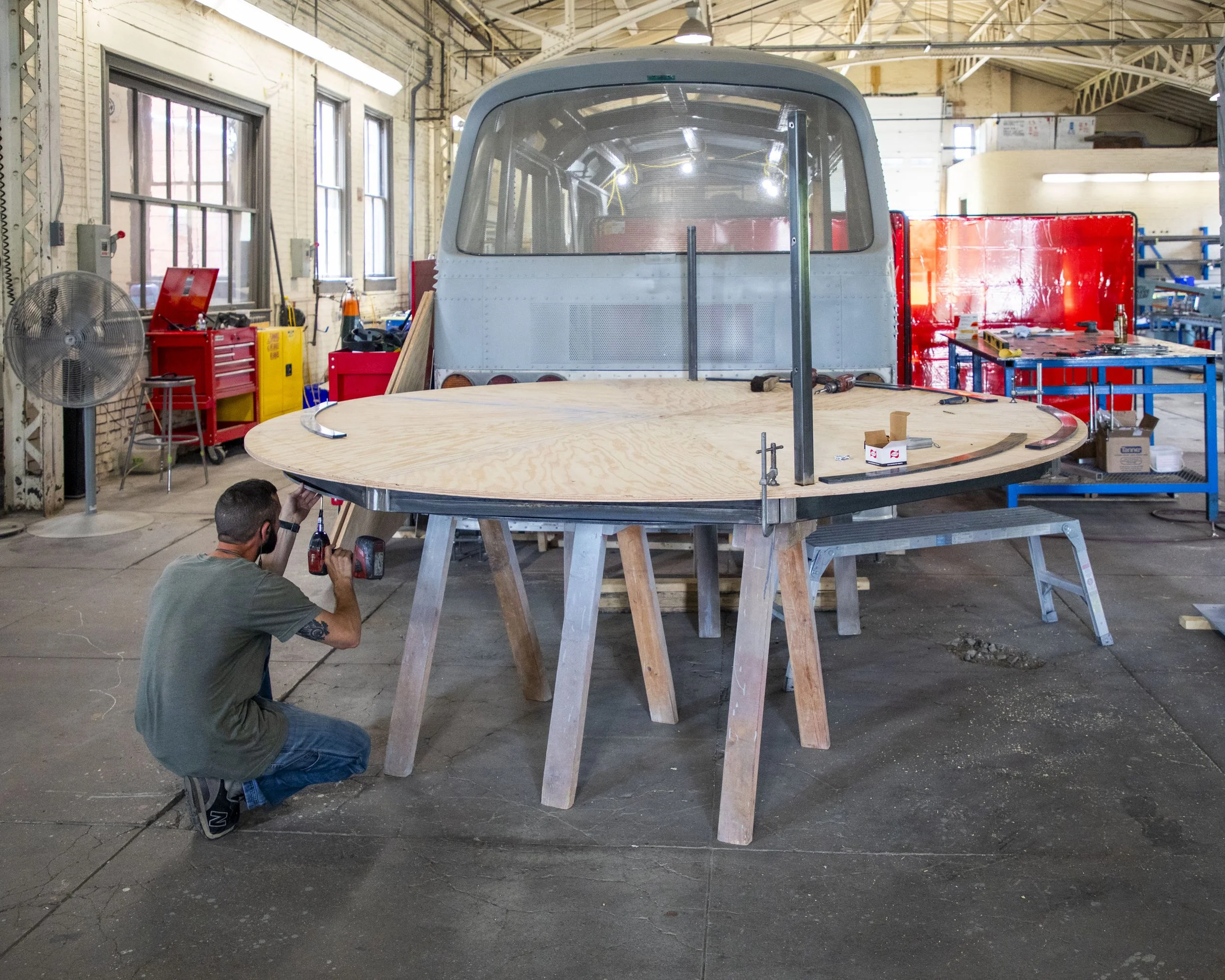 A person working on the construction of a boat's bow inside a workshop. The boat is elevated on wooden supports, with a large wooden deck and a partially installed cabin structure. Various tools and equipment are visible around the workshop.