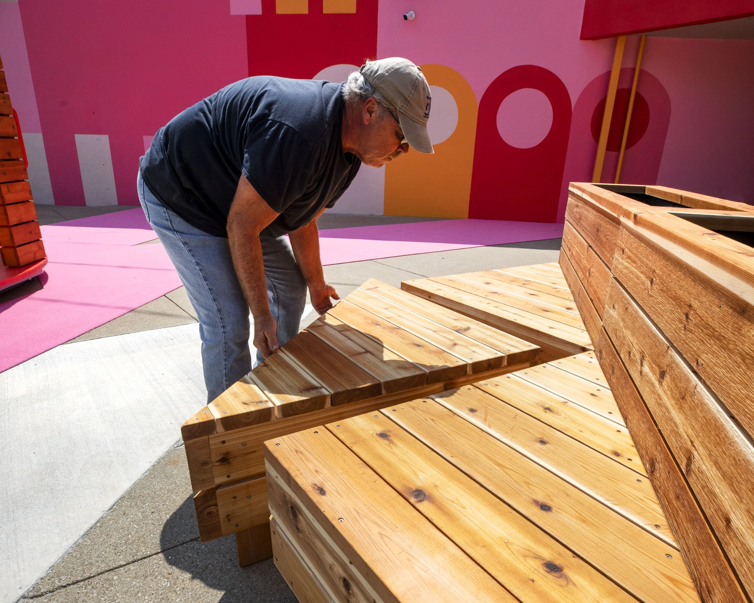 A man wearing a gray hat and black T-shirt bends over to adjust a wooden bench made of varnished planks, set against a colorful pink and orange mural background.