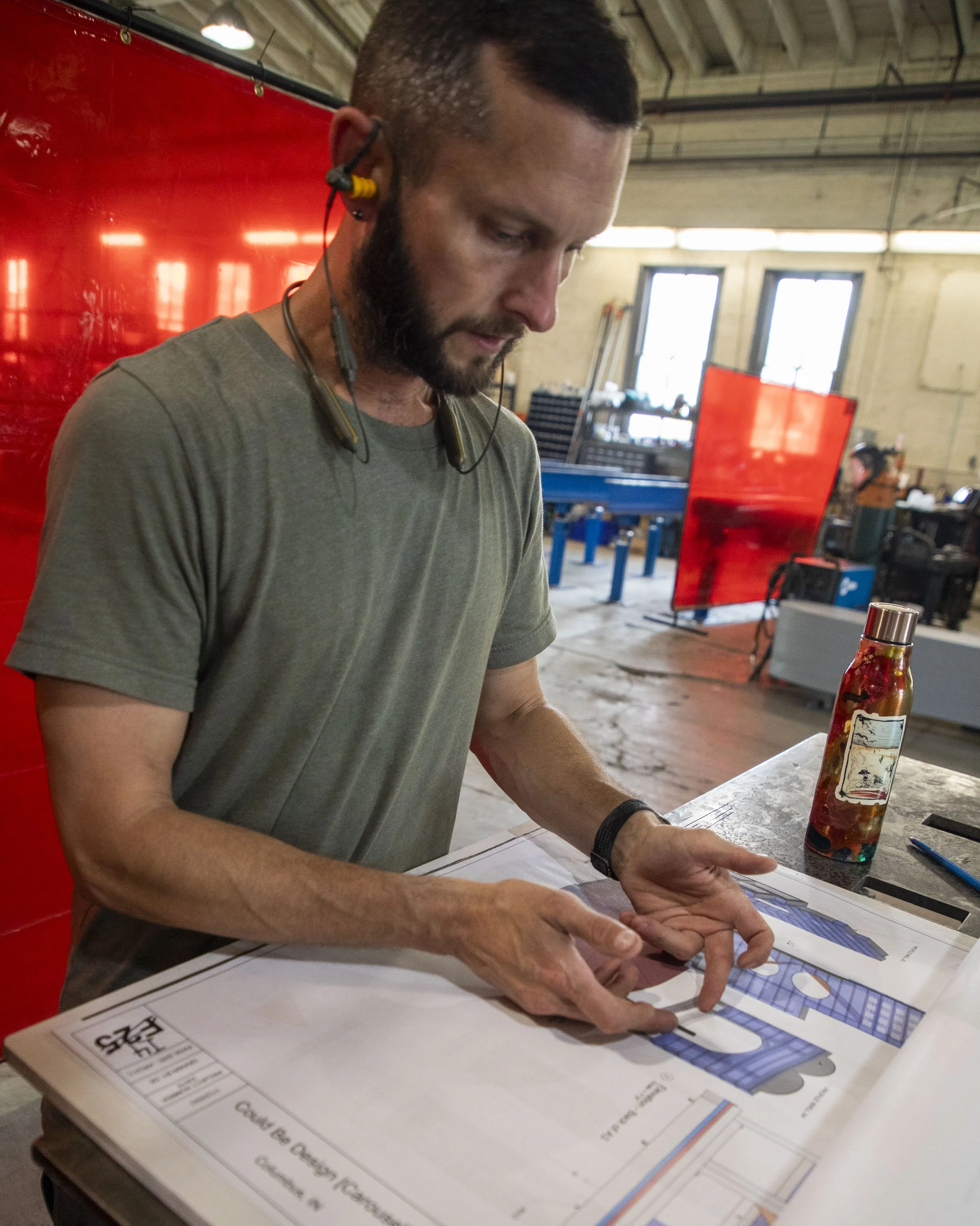 A man with a beard and wearing a gray t-shirt, with yellow and black ear protection, is looking down and pointing at a blueprint or technical drawing on a table in an industrial workshop. There is a water bottle and a pen on the table, and red welding screens are set up behind him.
