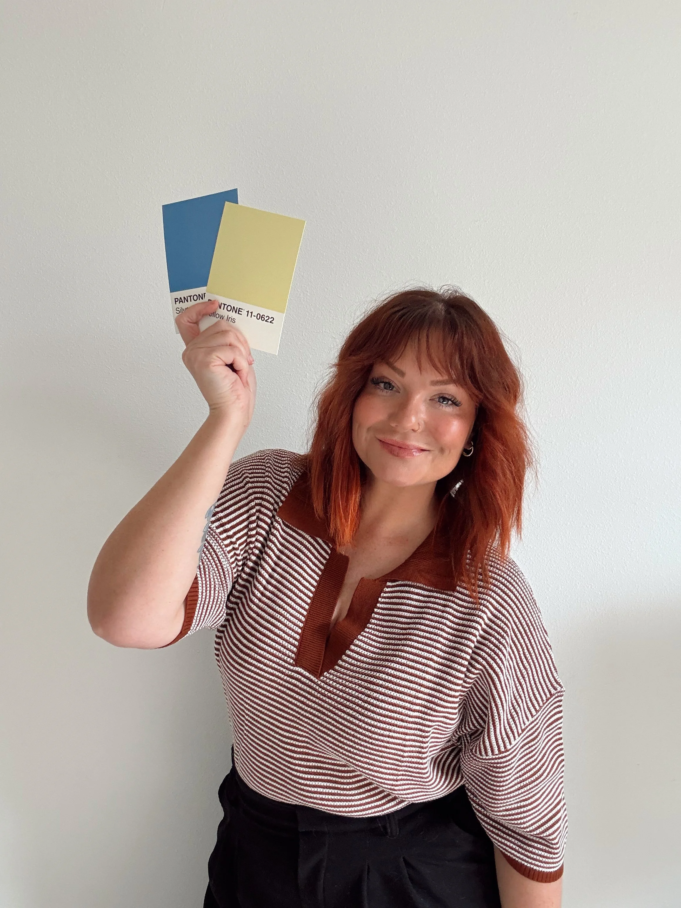 A woman with shoulder-length red hair and light skin, smiling, holding up two Pantone color swatches, blue and yellow, in front of a plain white wall.