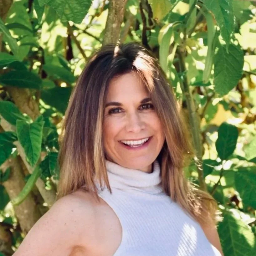 A woman with shoulder-length brown hair smiling outdoors among green plant leaves.