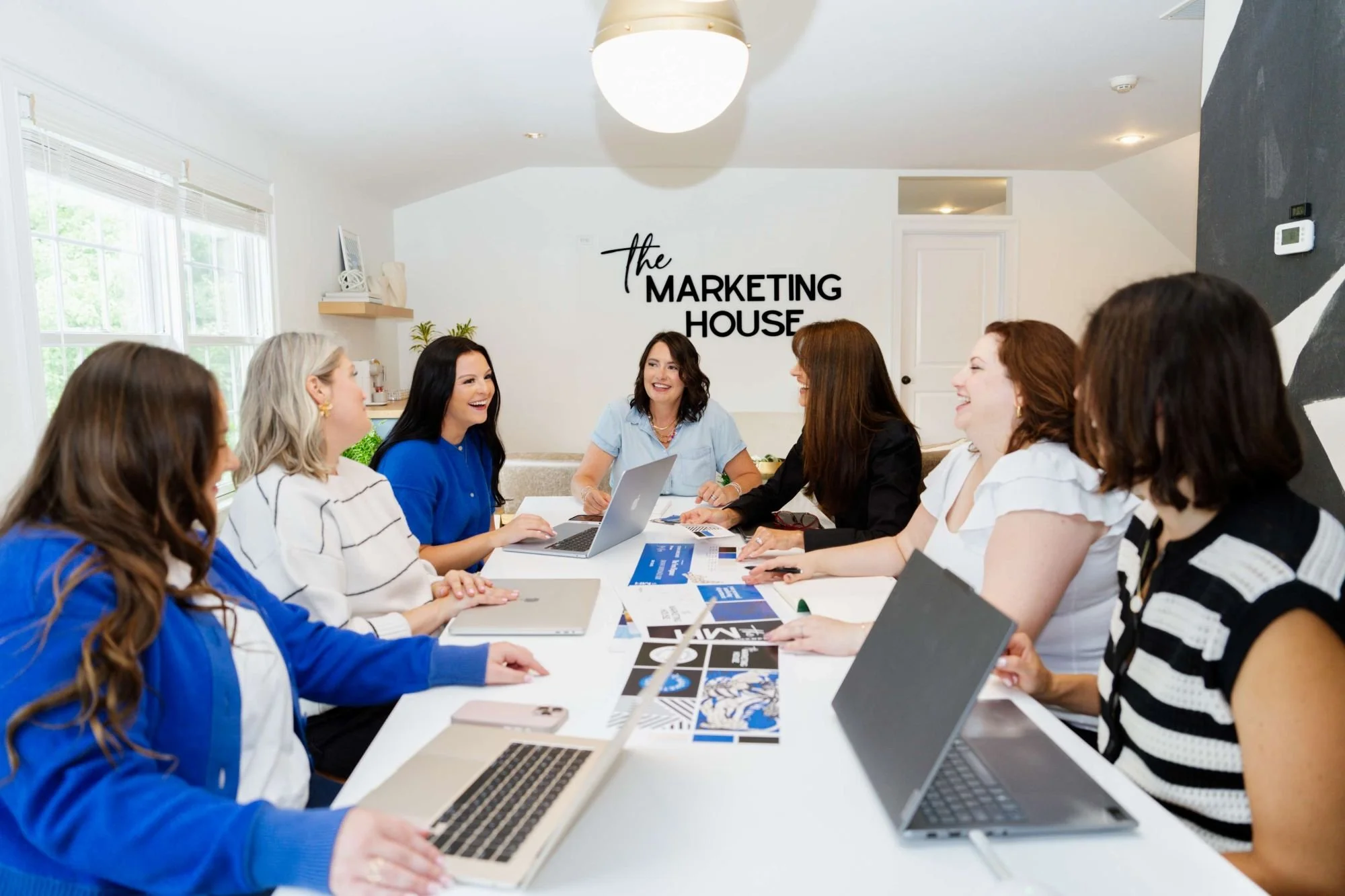 A group of women having a meeting in a conference room with windows, laptops, and printed materials, with a sign on the wall saying 'the MARKETING HOUSE'