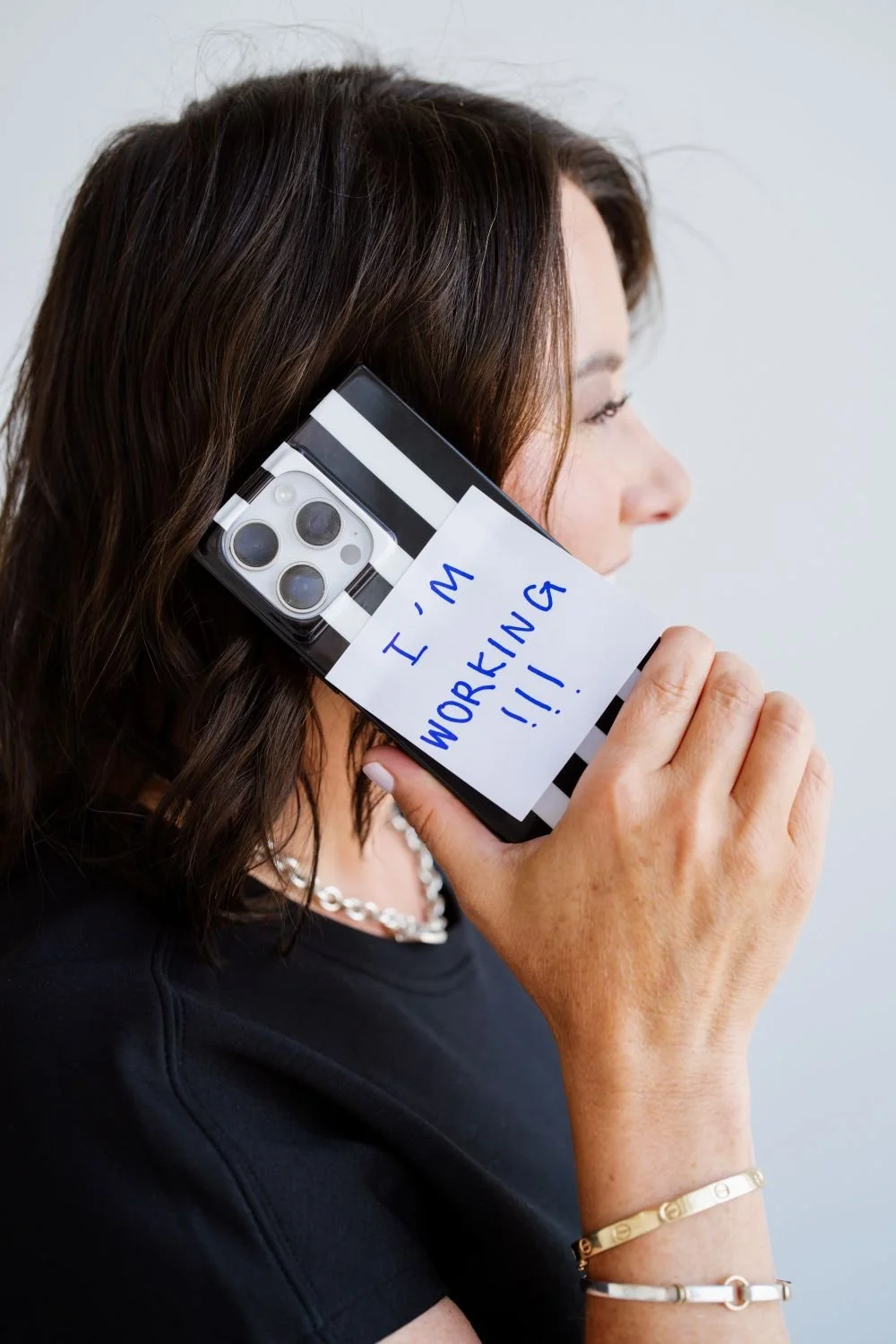 Woman holding a phone with a note attached that says 'I'm working!!!' over her ear.