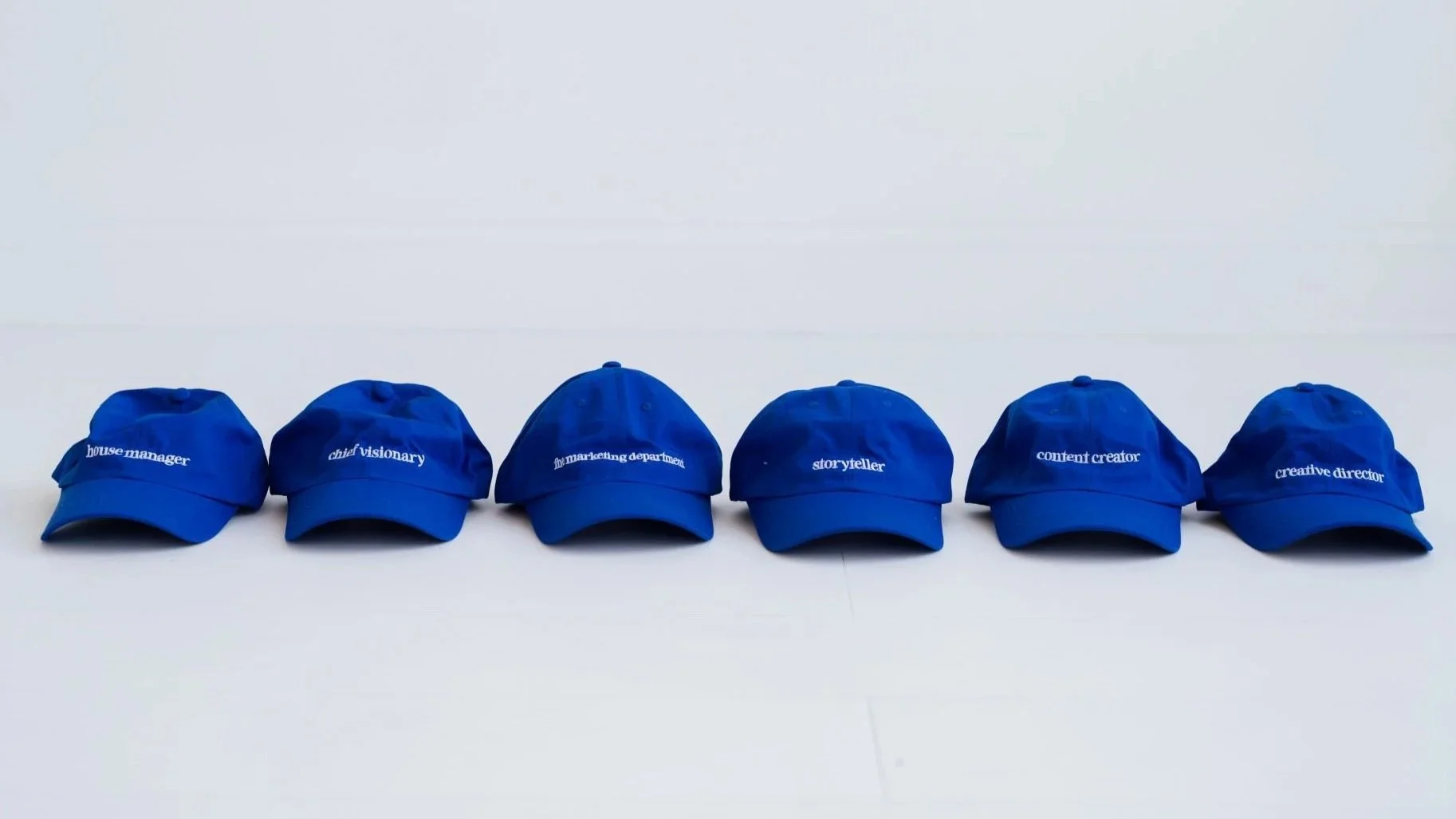 Six blue baseball caps with white embroidery displaying career titles, lined up on a white surface against a white background.