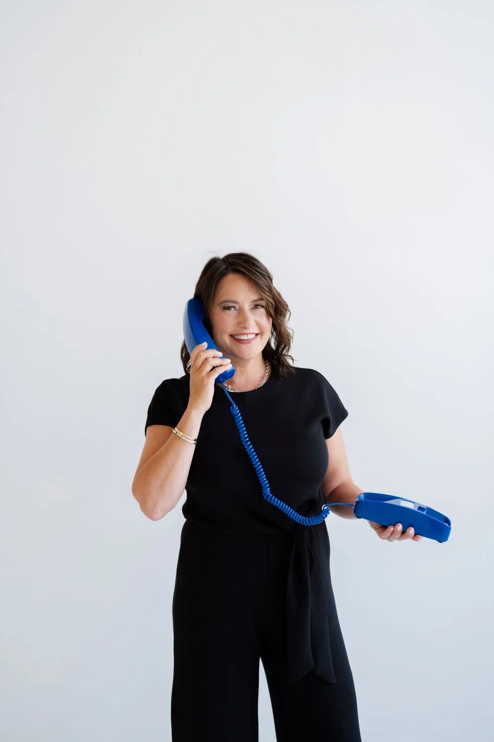 Smiling woman with shoulder-length brown hair, wearing a black outfit, holding a blue corded telephone to her ear, standing against a plain white background.