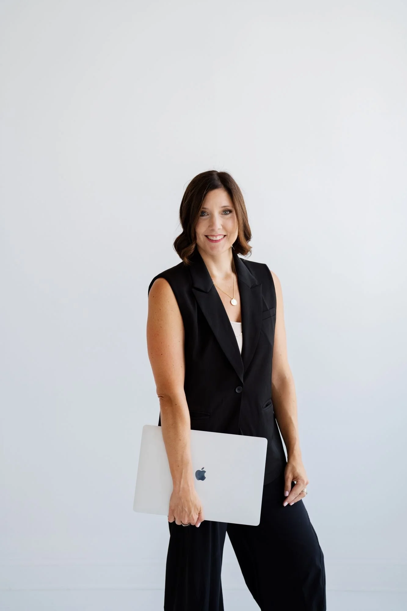 A woman with shoulder-length brown hair wearing a black sleeveless blazer, holding a silver MacBook in her left hand, standing in front of a plain white wall.