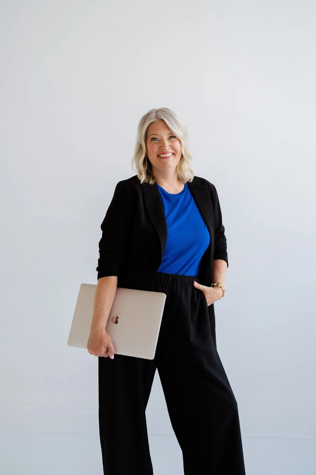 A woman with blonde hair wearing a black blazer and blue top, holding a silver MacBook, standing against a plain white wall.