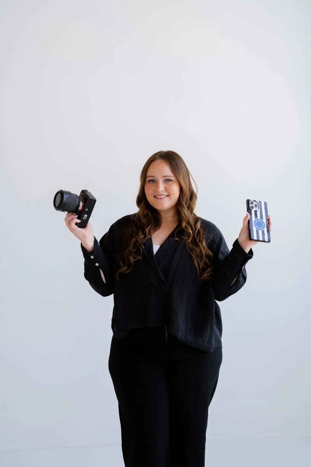 A woman with long curly hair standing against a plain light background, holding a camera in her right hand and a smartphone in her left hand, smiling at the camera.